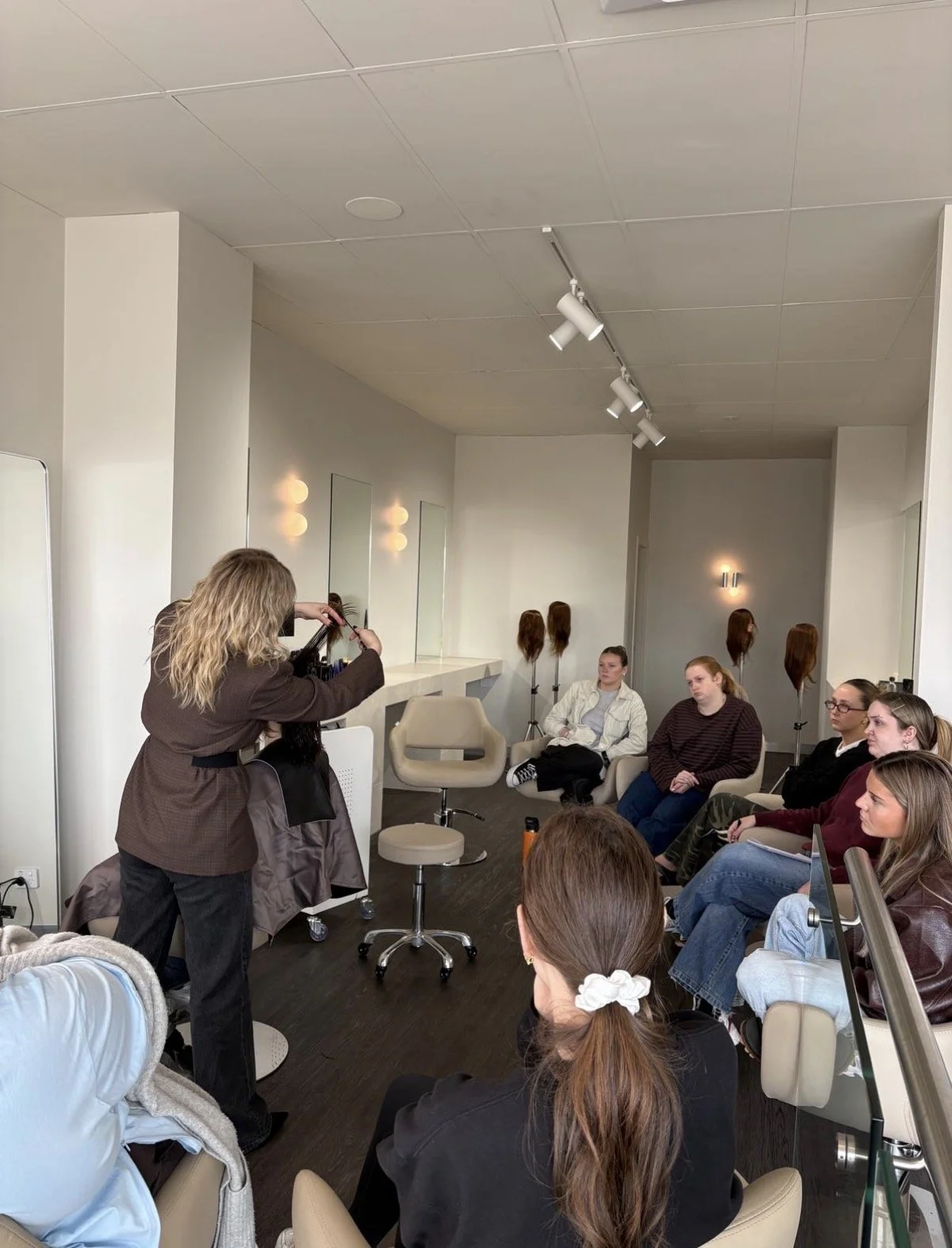 A hairstylist is demonstrating hair cutting techniques on a mannequin head to a group of women sitting in a salon or classroom setting. The room has white walls, mirrors, and wig heads in the background.