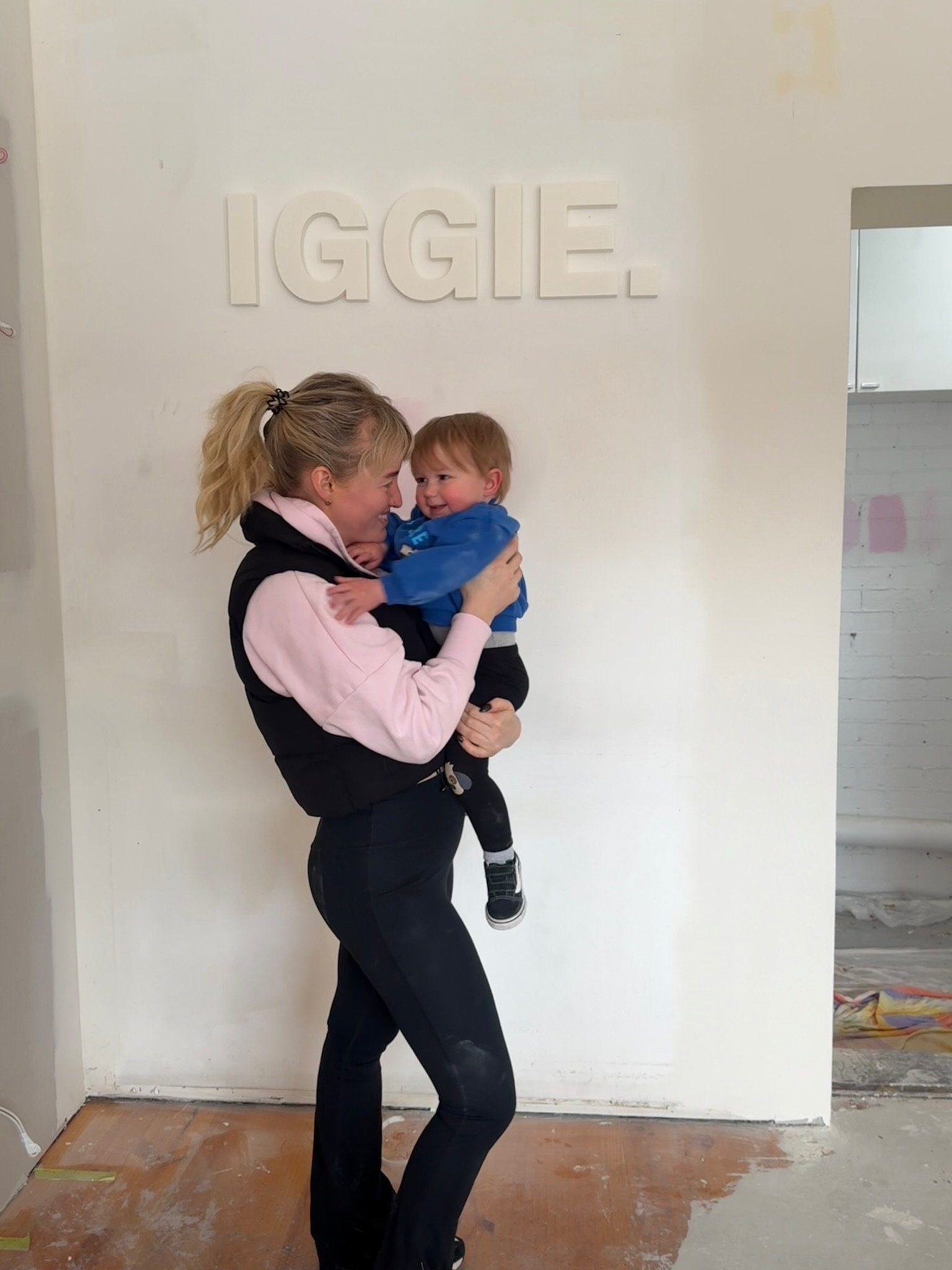 A woman with blonde hair in a ponytail holding a young boy with red hair and a blue jacket in front of a white wall with the word 'I G G I E' in raised white letters, during a home renovation.