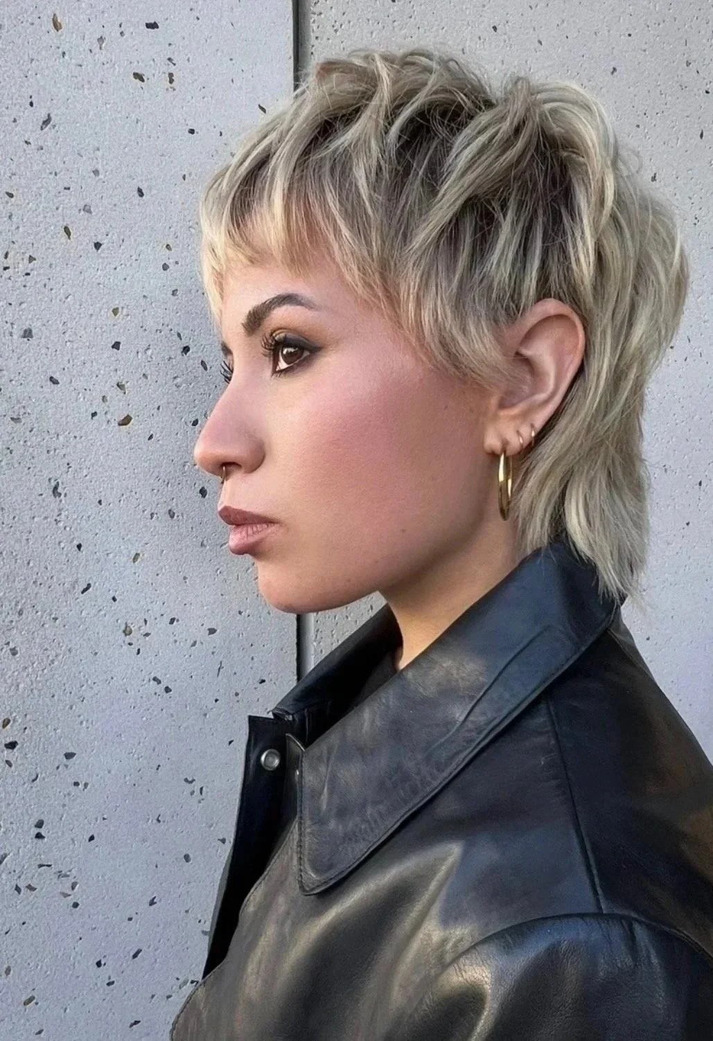 Profile of a woman with short, layered blonde hair, wearing hoop earrings and a black leather jacket, standing against a textured gray wall.