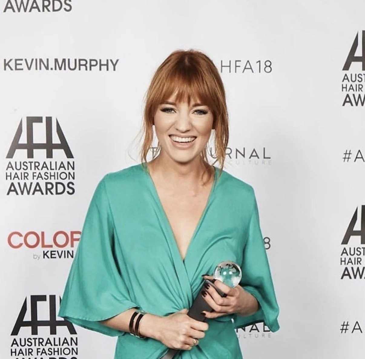 A woman with red hair, wearing a green dress, holding a glass trophy, smiling, at an awards event.