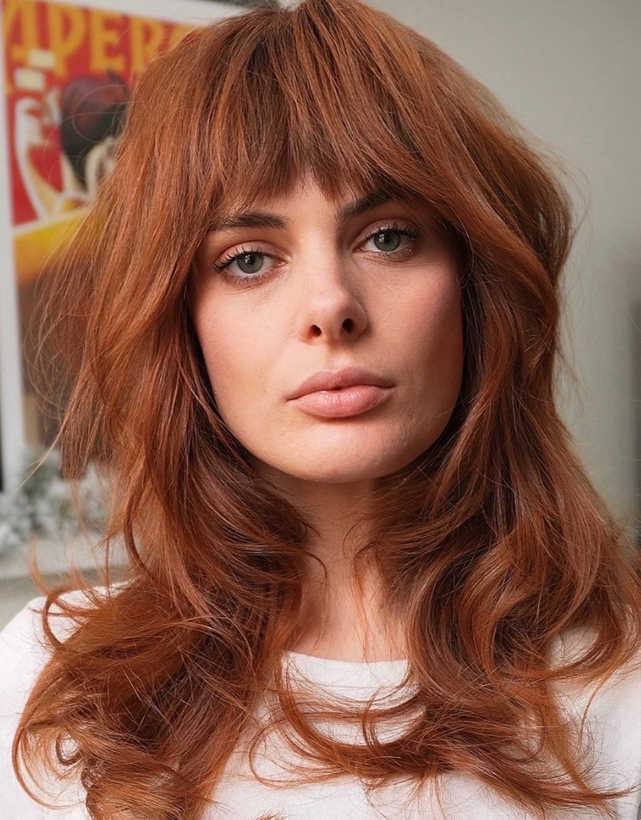 A woman with long, wavy red hair and gray eyes looks directly at the camera. She has a neutral expression and is wearing a light-colored top. In the background, there is a colorful poster.