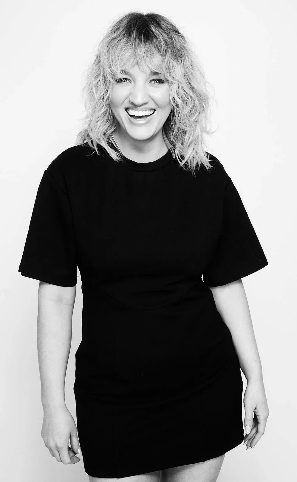 Black and white photo of a woman with curly blonde hair, smiling, wearing a black t-shirt and black skirt, standing against a white background.