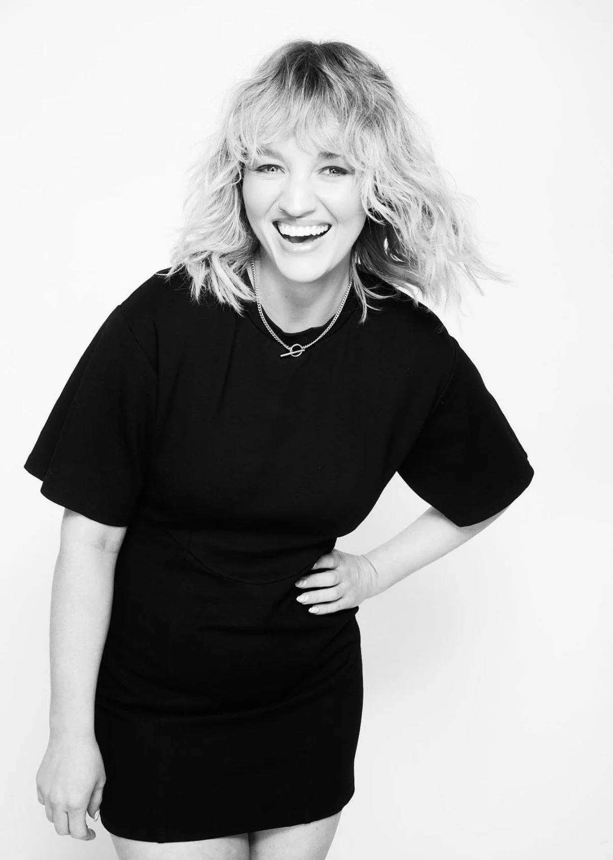 Black and white photo of a smiling woman with curly hair, wearing a black dress and jewelry, standing against a plain background.