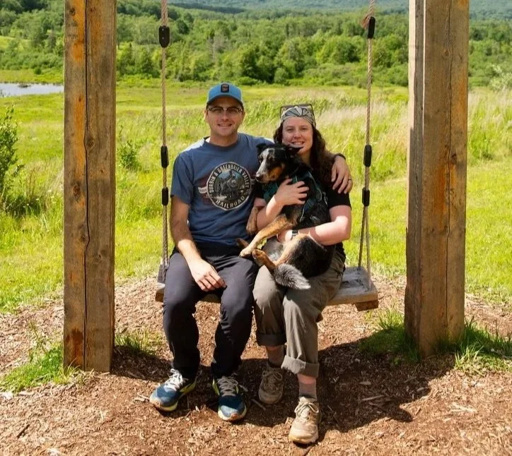 A man and woman sit on a wooden swing set outdoors in a grassy field with trees in the background, holding a black and brown dog between them, all smiling at the camera.