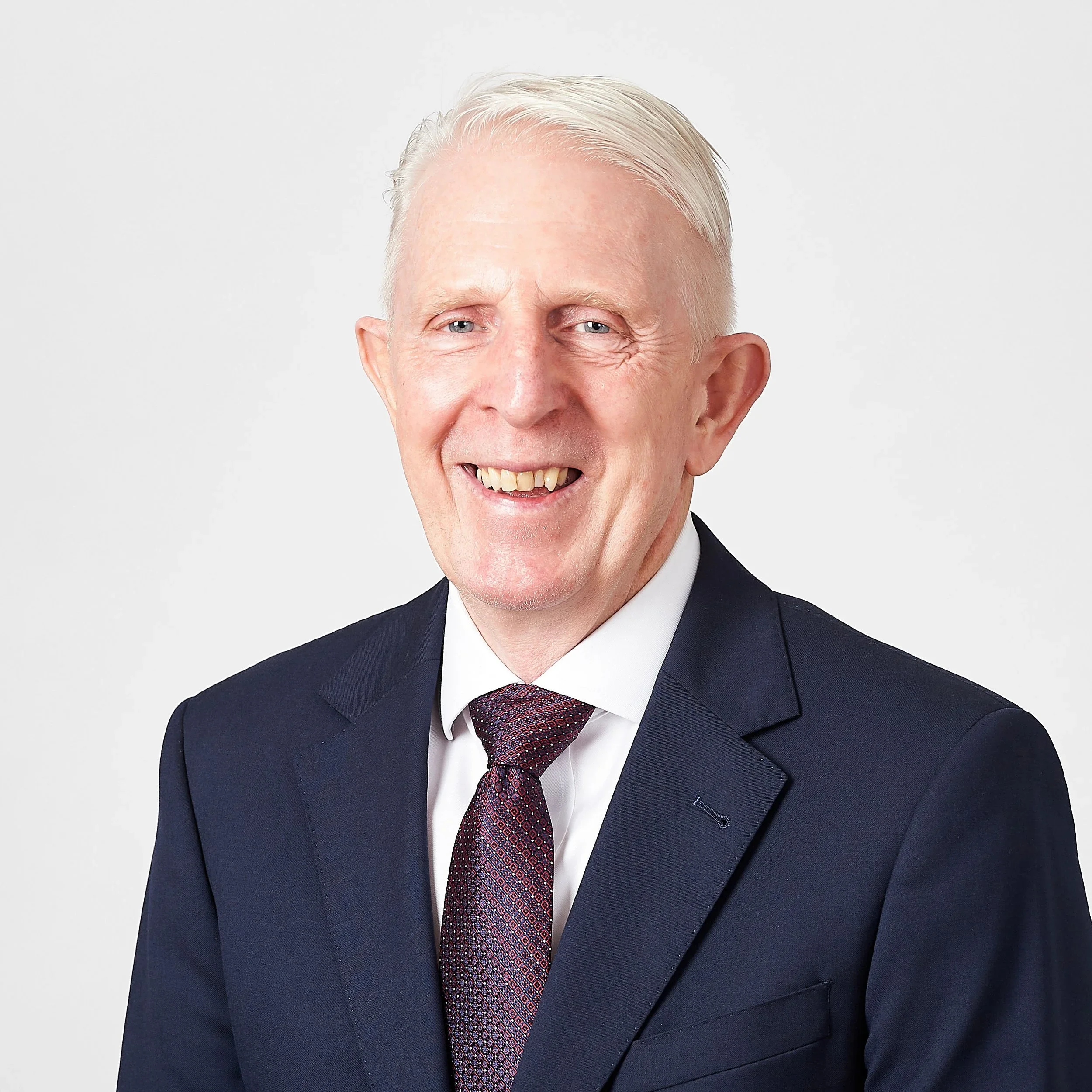 Close-up portrait of an older man with light hair, smiling, wearing a dark suit, tie, and white shirt, against a plain white background.