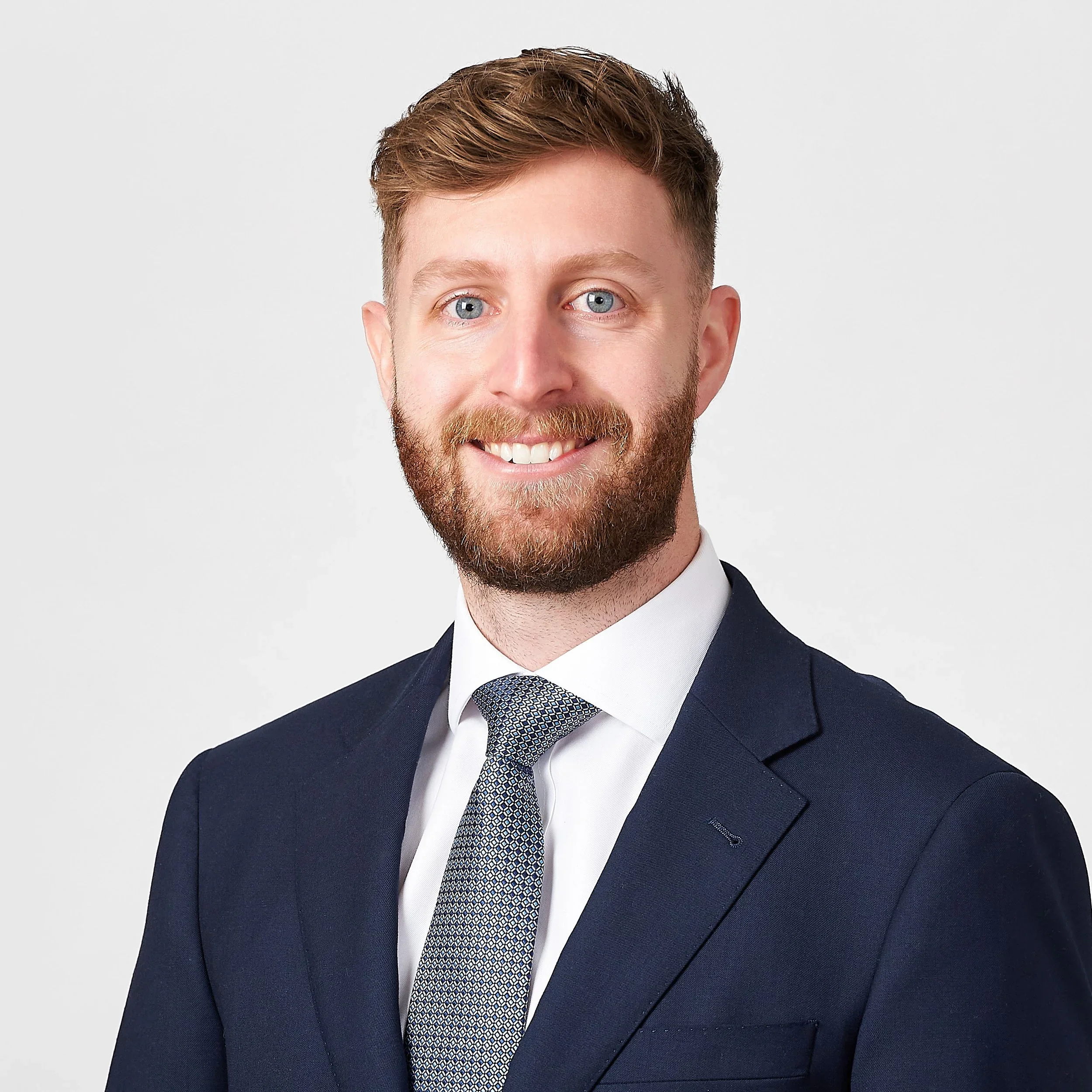 Close-up of a young man with reddish-brown hair, blue eyes, and a beard, smiling, wearing a navy suit, white shirt, and a patterned tie, against a plain white background.