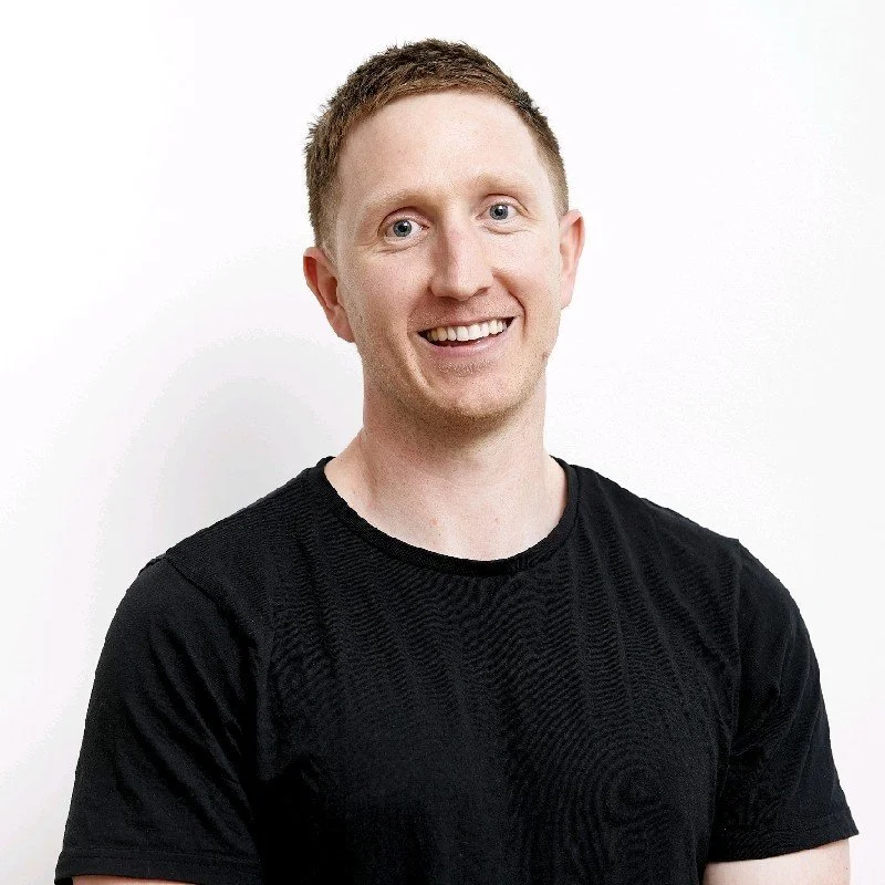 A smiling man with short red hair, wearing a black t-shirt, standing against a white background.
