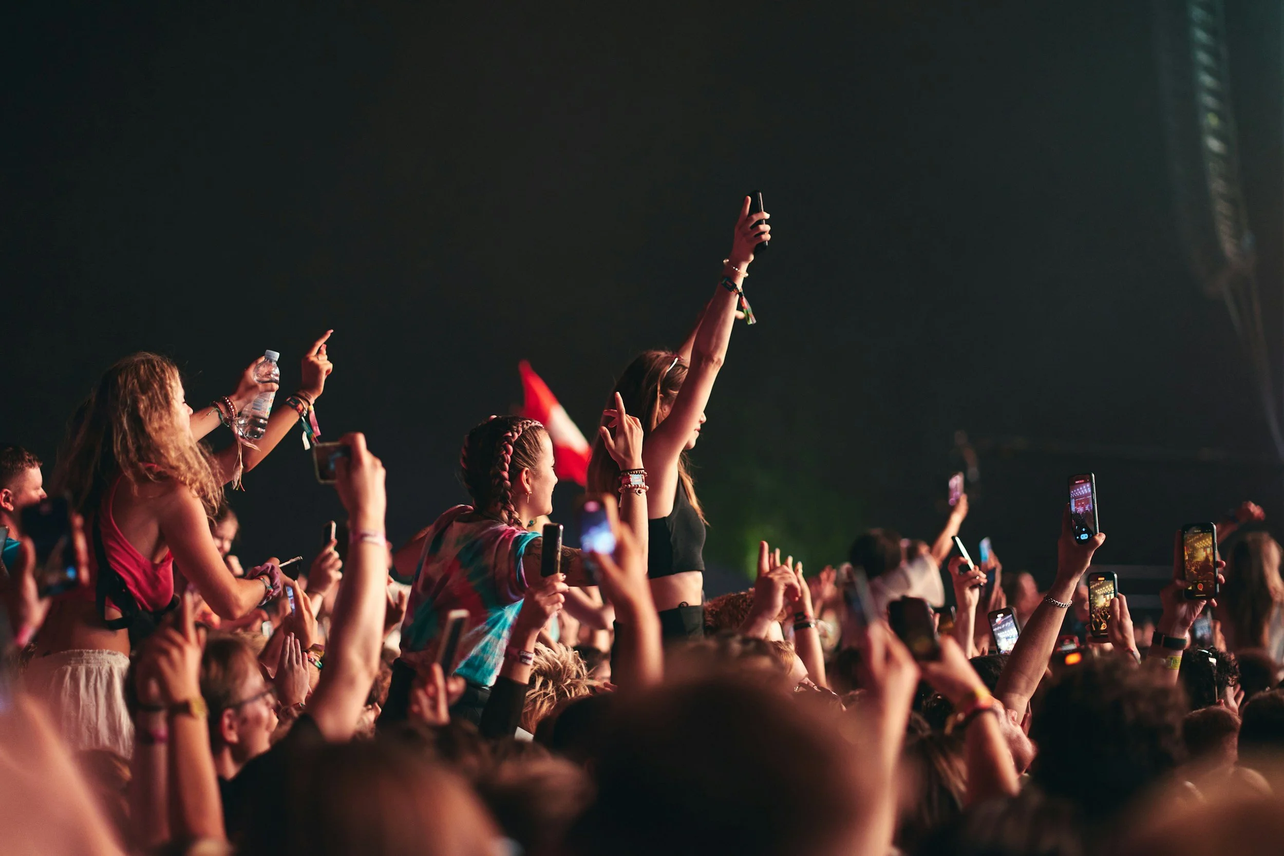 Concert crowd with people holding up smartphones, some raising hands, at night with dark background.