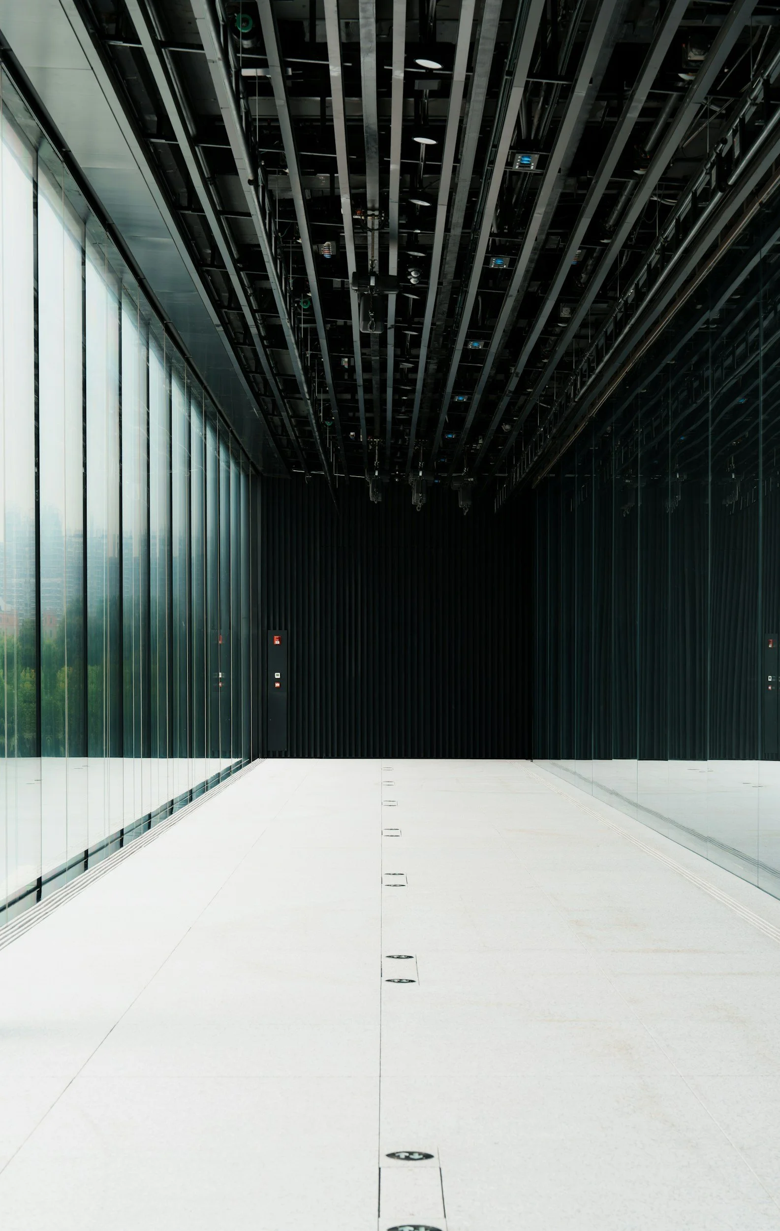 Modern interior space with white tiled floor, glass wall on the left, black wall on the right, and an exposed ceiling with visible metal beams and equipment.