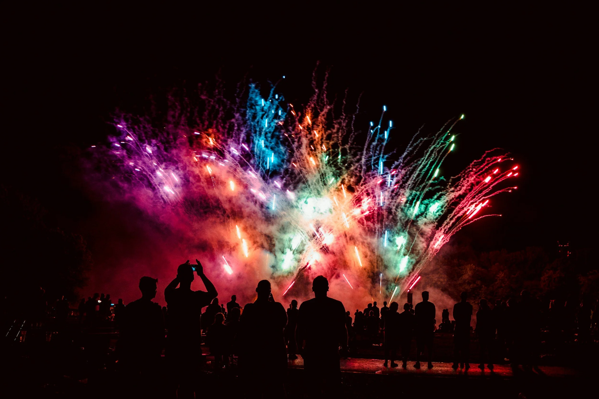 People watching colorful fireworks display at night with silhouettes capturing photos.