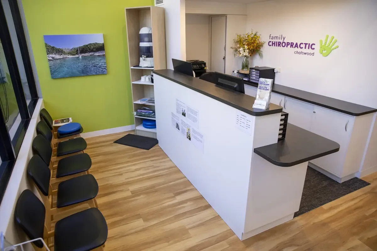 Reception area of a chiropractic office with black chairs, a green wall, a water fountain, a camera, a bookshelf, and a desk with a computer, brochures, and flowers. The wall displays the logo 'family CHIROPRACTIC chatswood' with a green hand and leaf design.