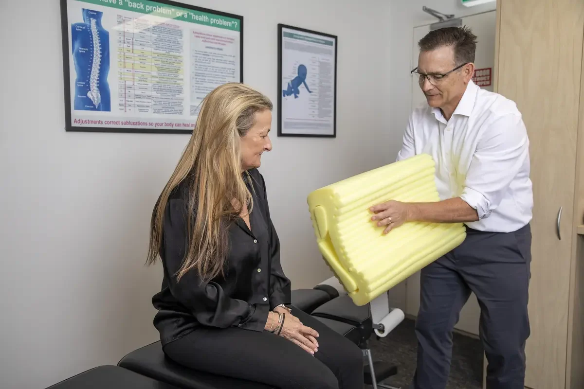 Dr Brett Grant, chiropractor holding a yellow foam roller in front of a woman seated on an examination table in a clinic