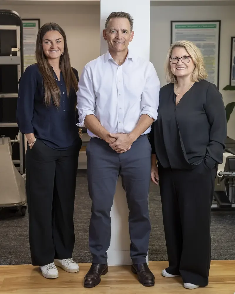 Three adults, two women and one man, standing indoors in front of a white column and smiling at the camera. The woman on the left has long brown hair, is wearing a navy blouse and black pants. The man in the middle has short hair, is wearing a white dress shirt and gray pants. The woman on the right has blonde hair, glasses, and is wearing a dark blazer and pants.