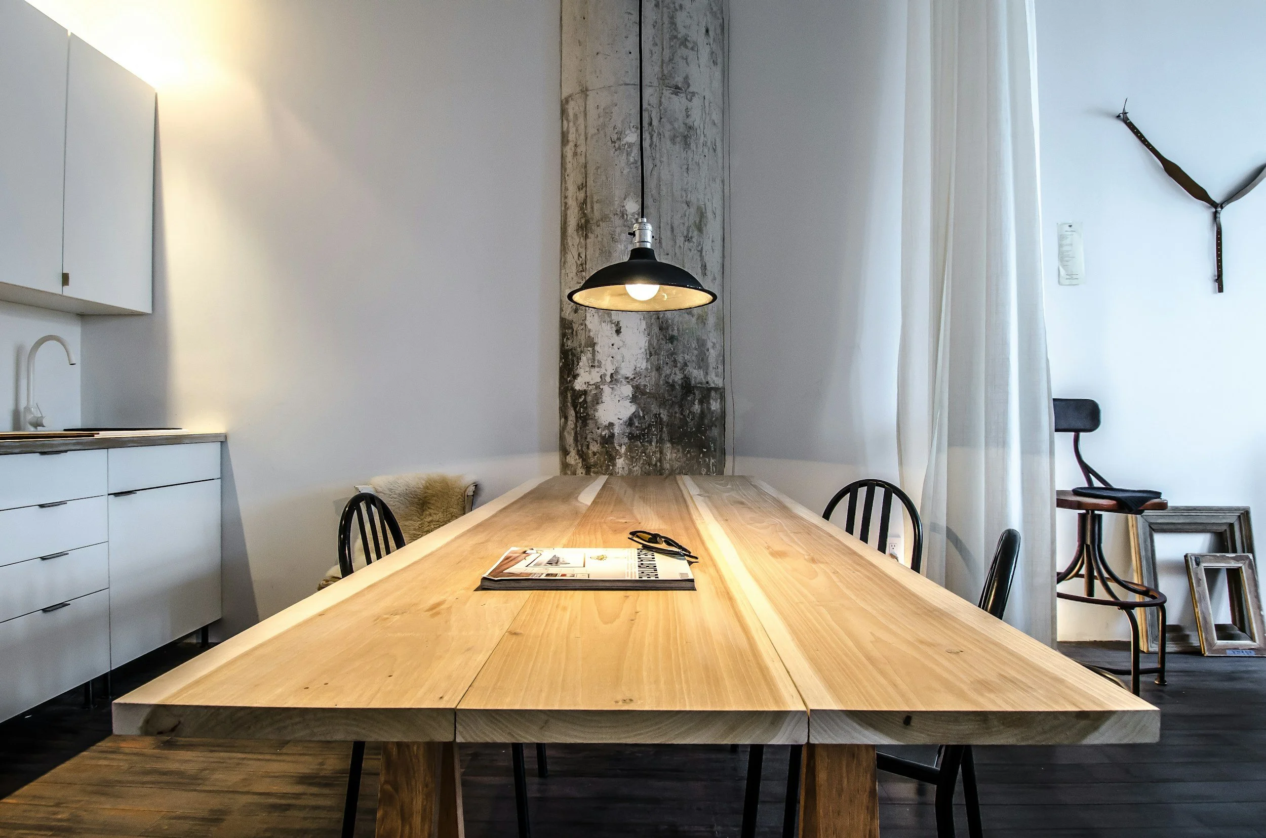 A modern dining area with a large wooden table, black chairs, a pendant light hanging above the table, a white cabinet with drawers on the left, a concrete accent wall behind the table, a sheer white curtain on the right, and various framed pictures on the floor to the right.