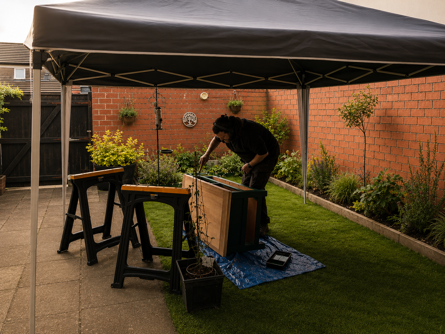 A person working on a gardening project under a black canopy in a backyard, with tools and plants around.
