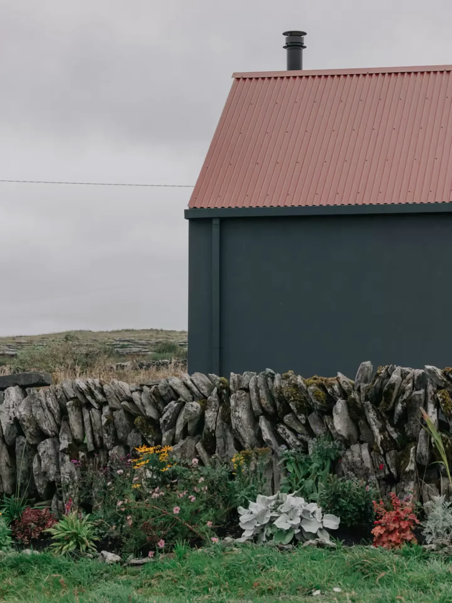 A house with a red metallic sloped roof, dark green walls, a stone fence, and a garden with various colorful flowers in front under an overcast cloudy sky.