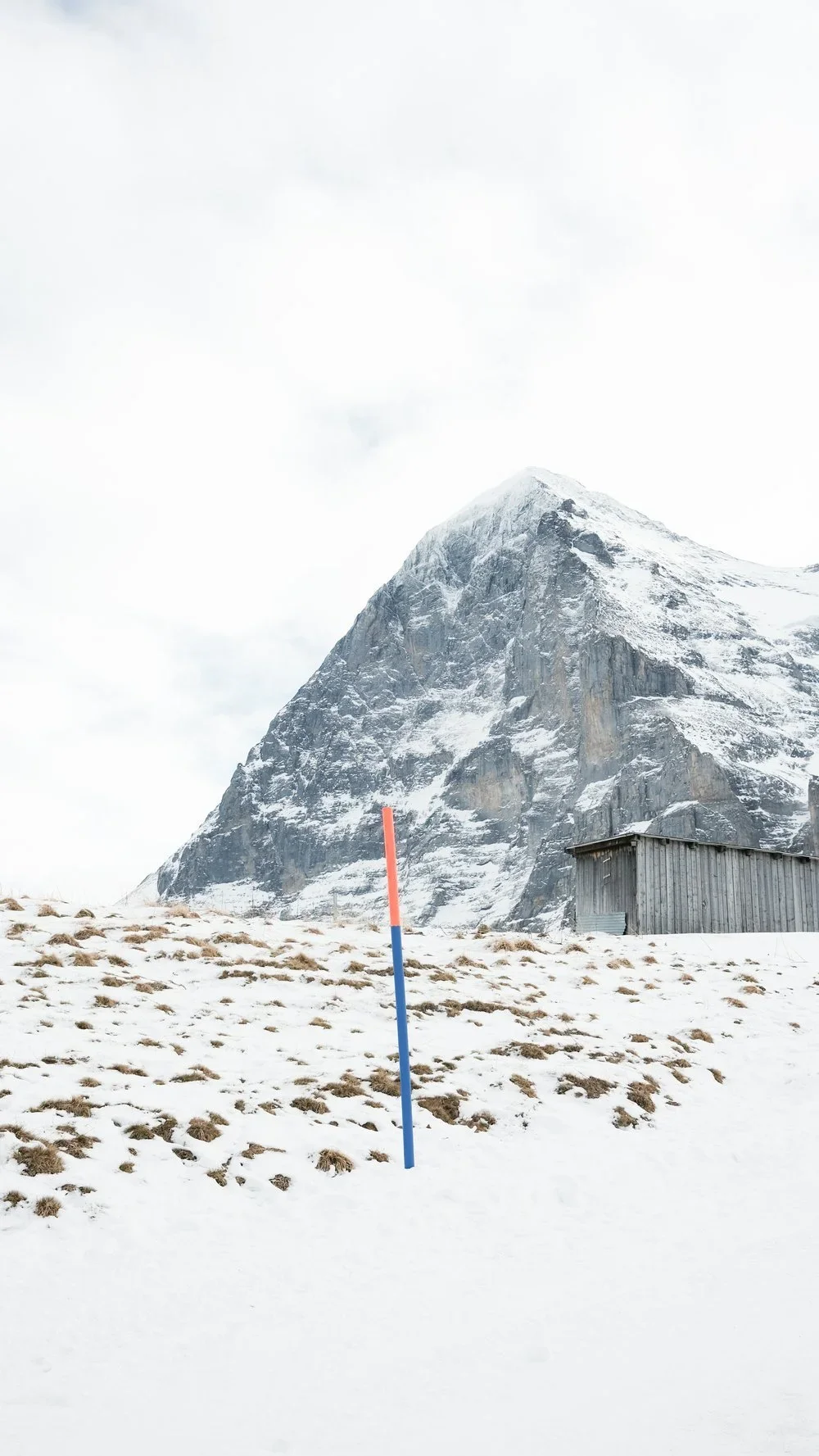 Snow-covered mountain with a small wooden shed nearby, and a blue and red pole in the foreground.