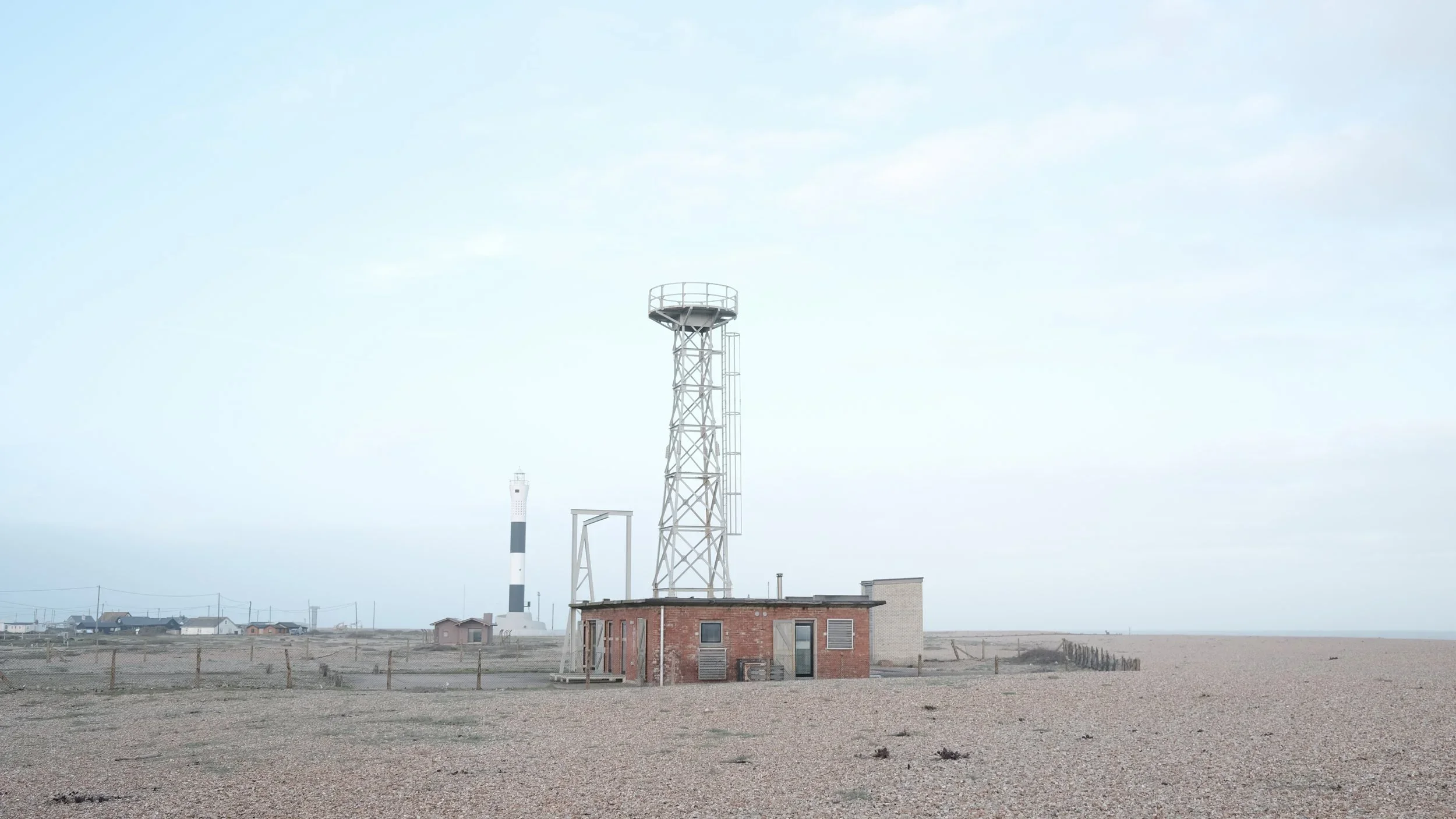 A deserted coastal area with a small brick building topped with a tall metal tower, and a lighthouse in the background under a cloudy sky.