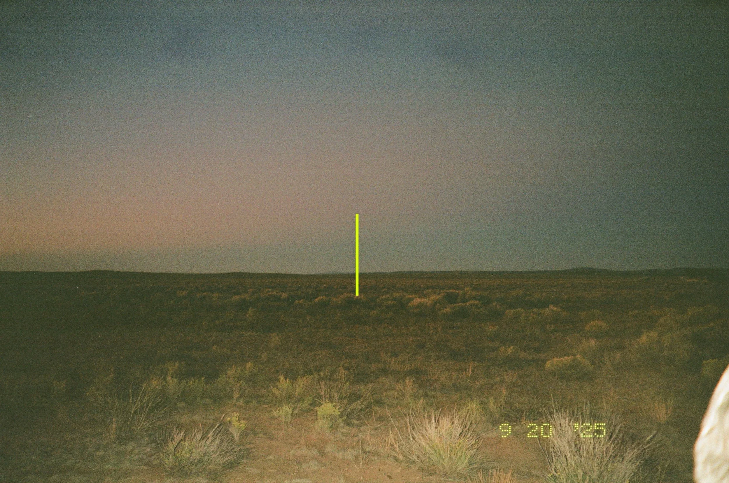 Nighttime landscape of a flat desert or plains with sparse vegetation under a clear sky, with a vertical green line in the center of the image.