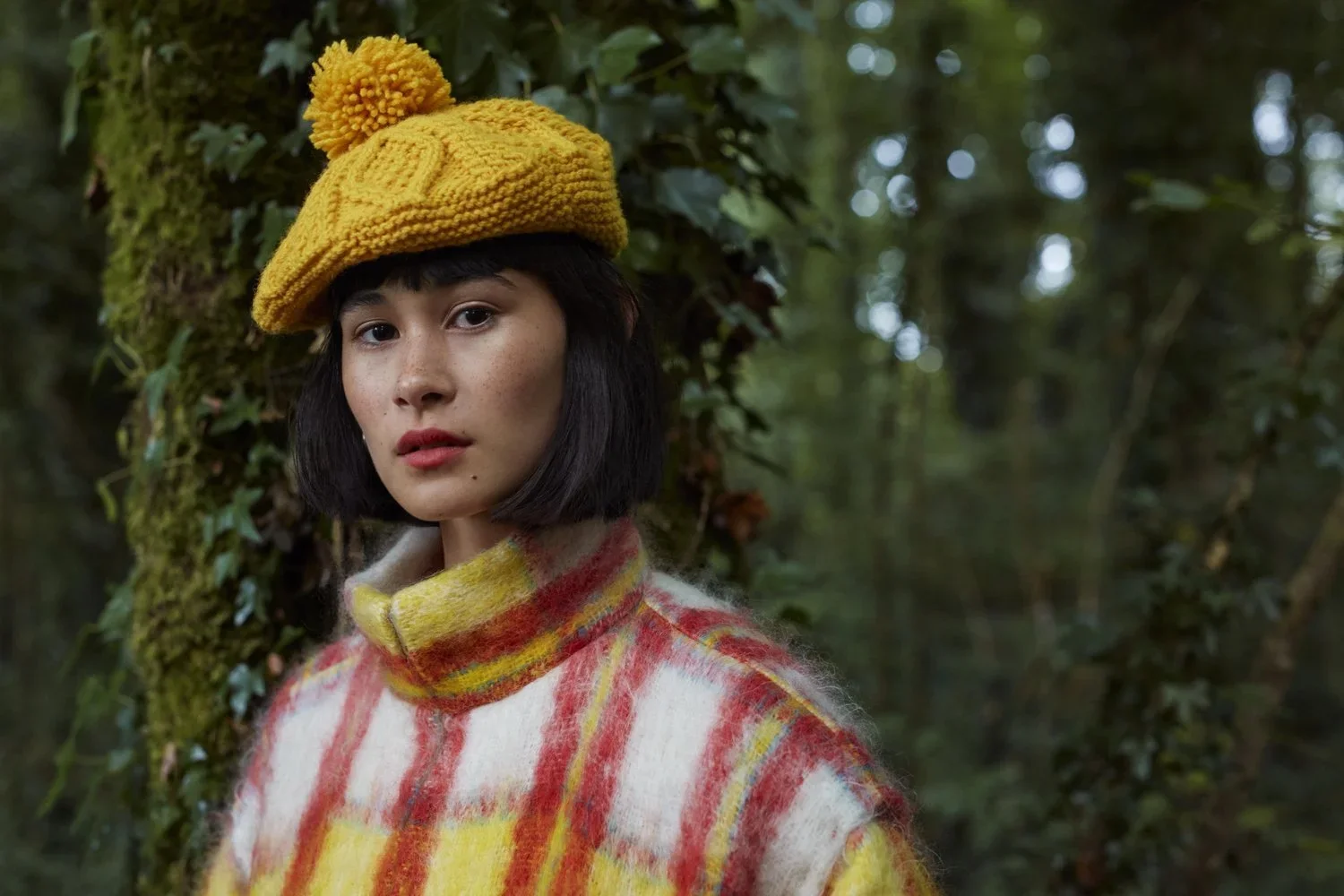 Young woman with short dark hair wearing a yellow knit hat with a pom-pom and a red, yellow, and white plaid sweater standing in a forest.