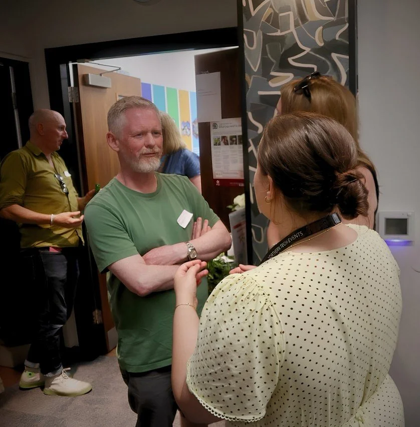 A group of people talking at an indoor event, with a man in a green shirt in the foreground and women facing him, one in a yellow polka-dot dress. A man in a yellow shirt stands in the background near the door.