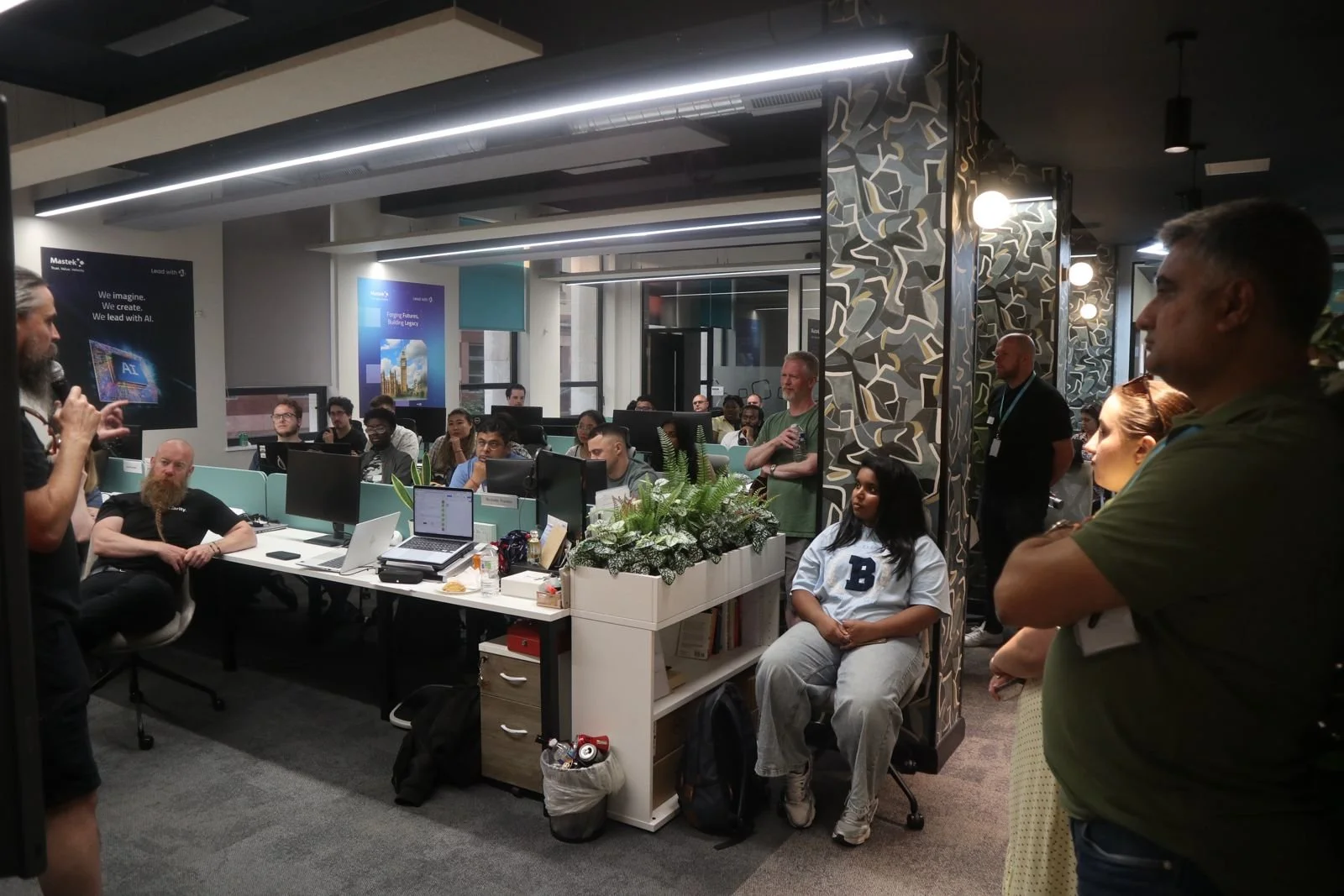 A group of people attending a presentation or meeting in an office, with a speaker on the left, some sitting at desks with computers, and others standing or listening.