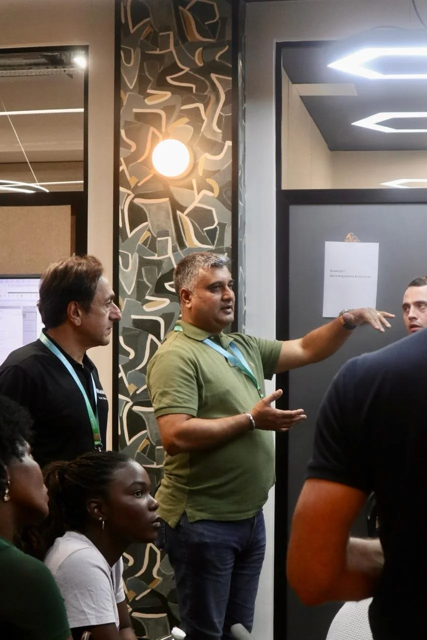 A man in a green polo shirt is speaking and gesturing with his right hand during a discussion or presentation. Three other people are listening attentively—two men standing on his left, and two women seated in the foreground. The background features a modern, abstract patterned wall with a round light fixture.