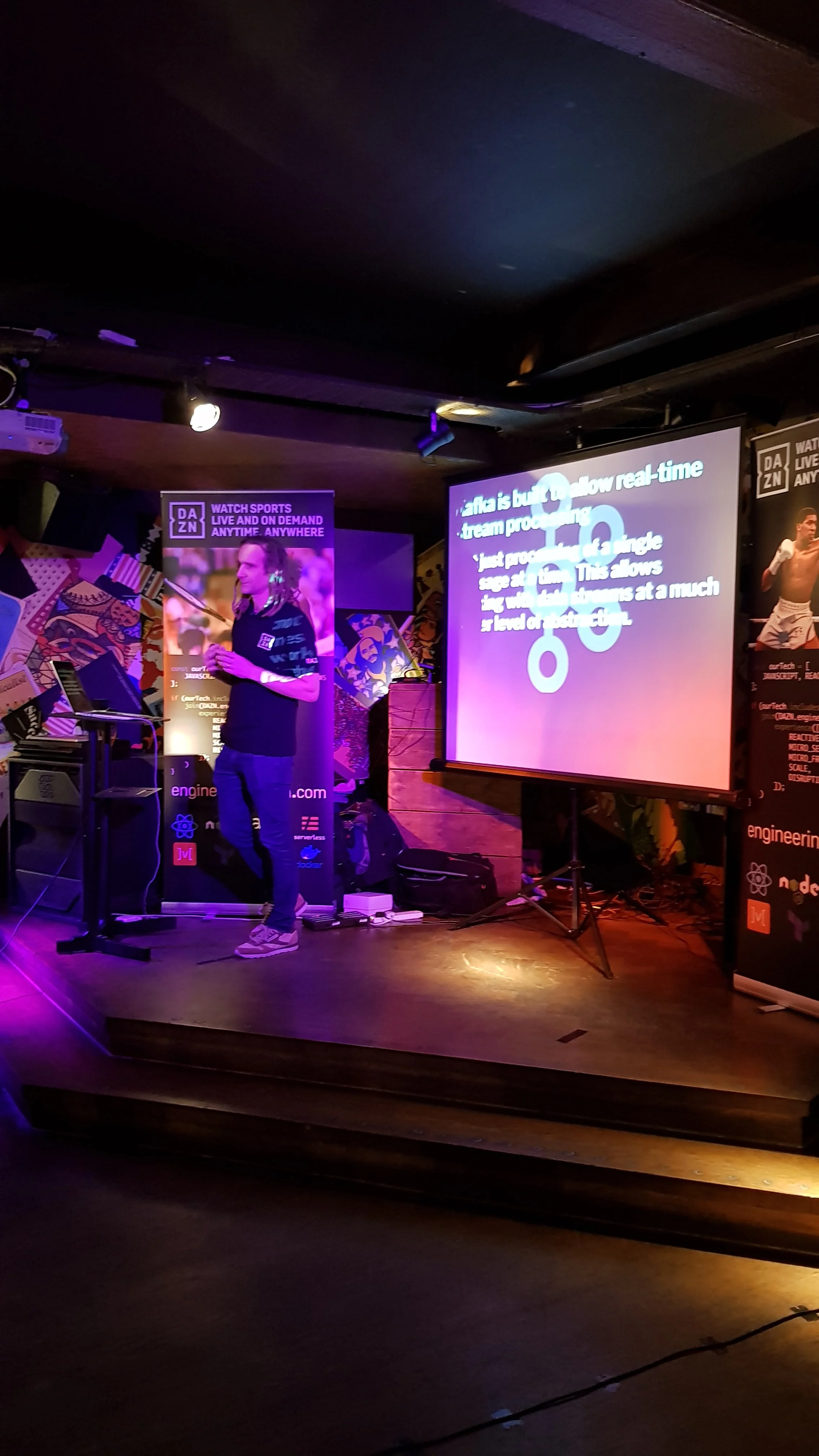 A man standing on a stage with a microphone, wearing a black shirt and jeans, giving a presentation with a large screen displaying a slide in the background. There are promotional banners for DAZN and various tech companies.