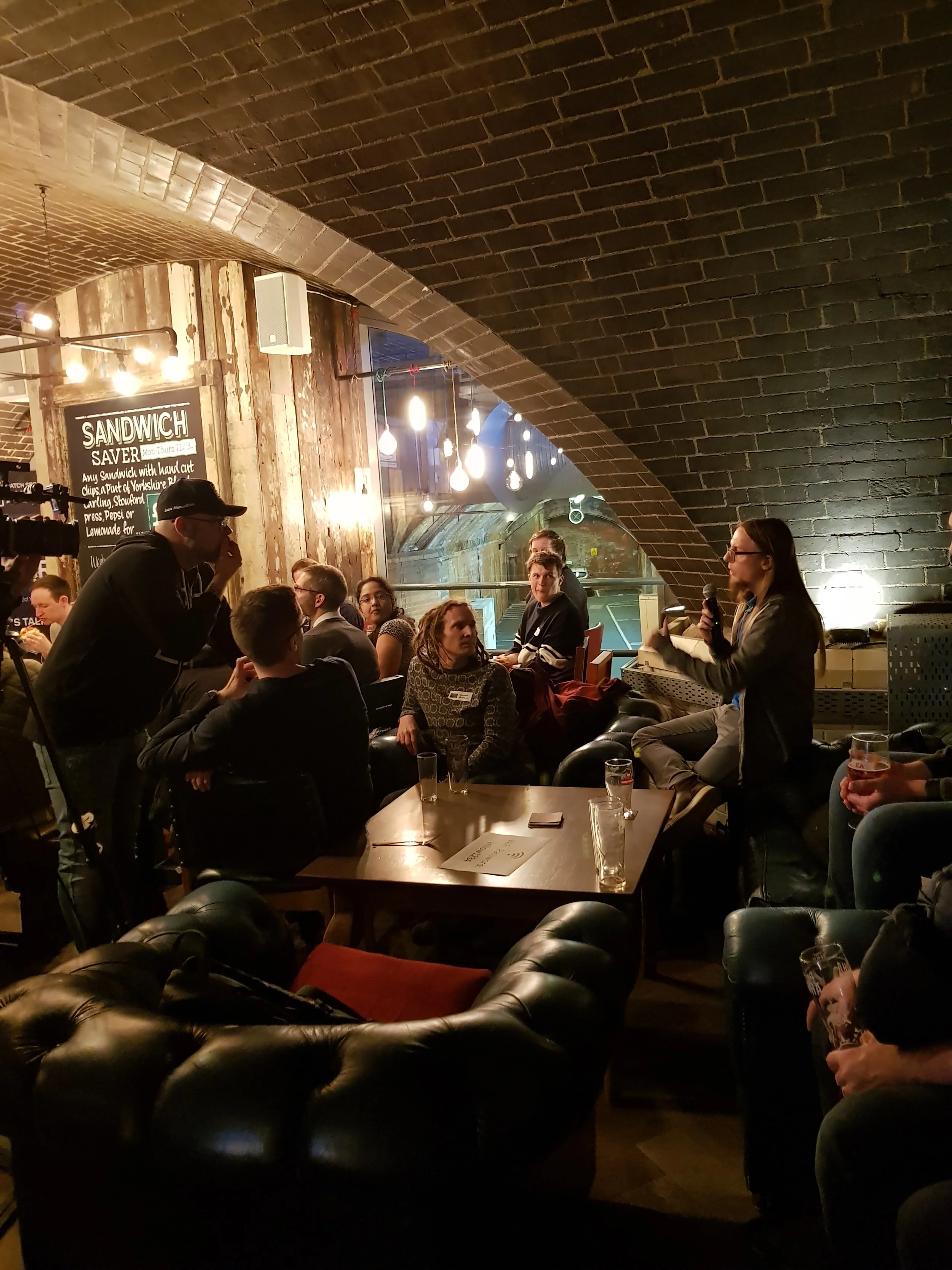 People gathered in a restaurant or bar beneath a brick arch ceiling, some sitting and some standing, with one woman speaking into a microphone, others listening, while drinks and glasses are on the tables.