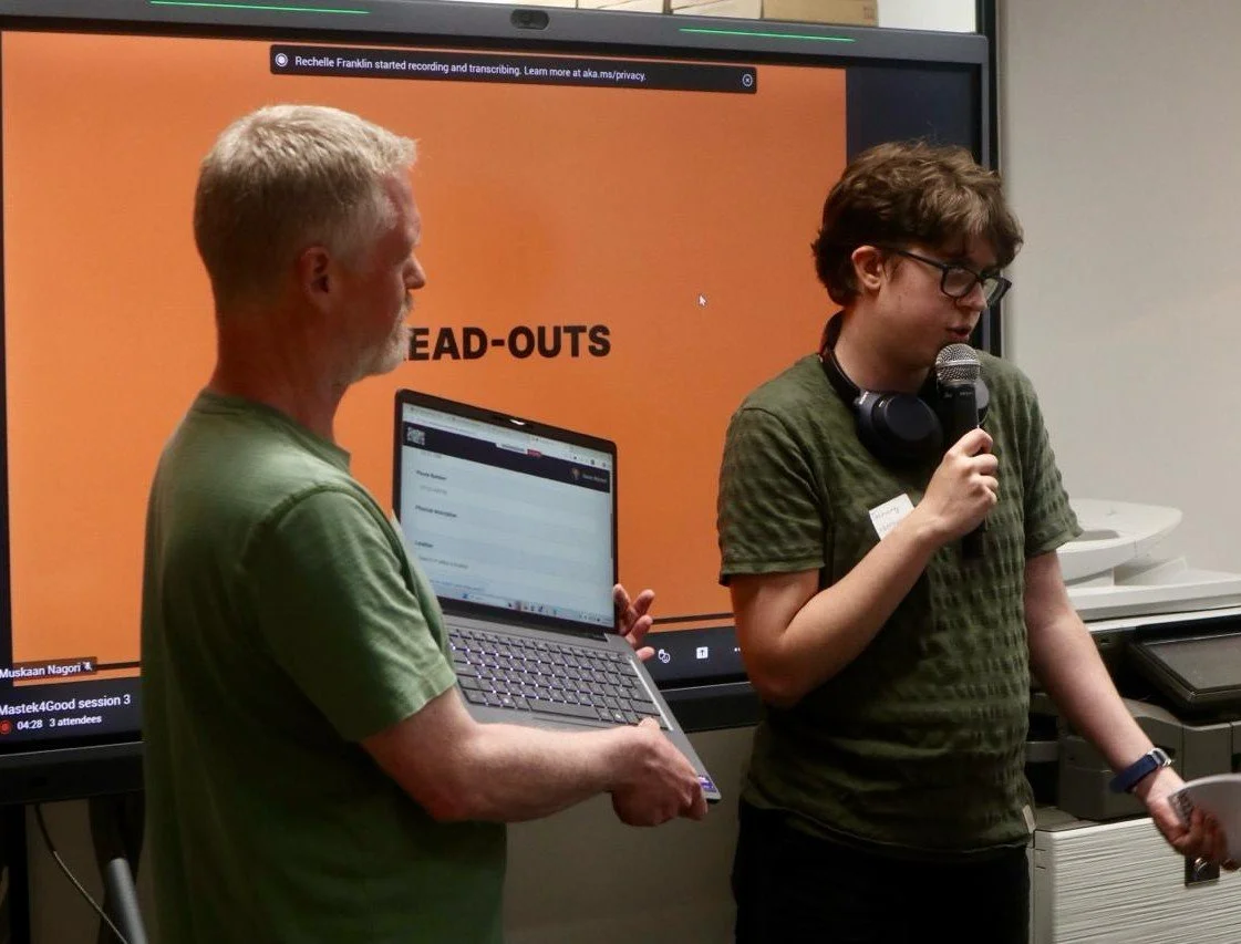 Two men speaking at a presentation, one holding a laptop and the other speaking into a microphone, in front of a screen with 'HEAD-OUTS' on an orange background.
