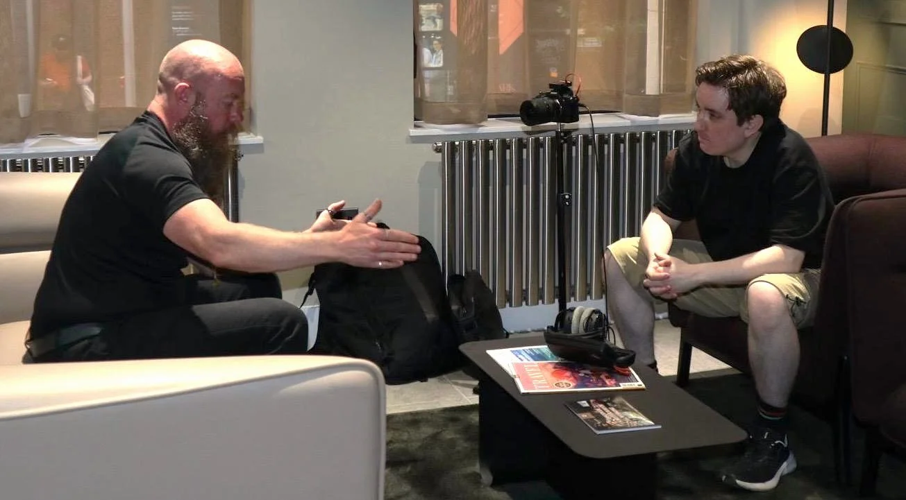 Two men are sitting across from each other in a hotel lobby or lounge area. The man on the left has a bald head, a full beard, and is wearing a black T-shirt. He is gesturing with his hands while talking. The man on the right has short, curly hair and is wearing a black shirt and khaki shorts, listening attentively. There are magazines, headphones, a camera, a backpack, and a remote control on a small table between them.