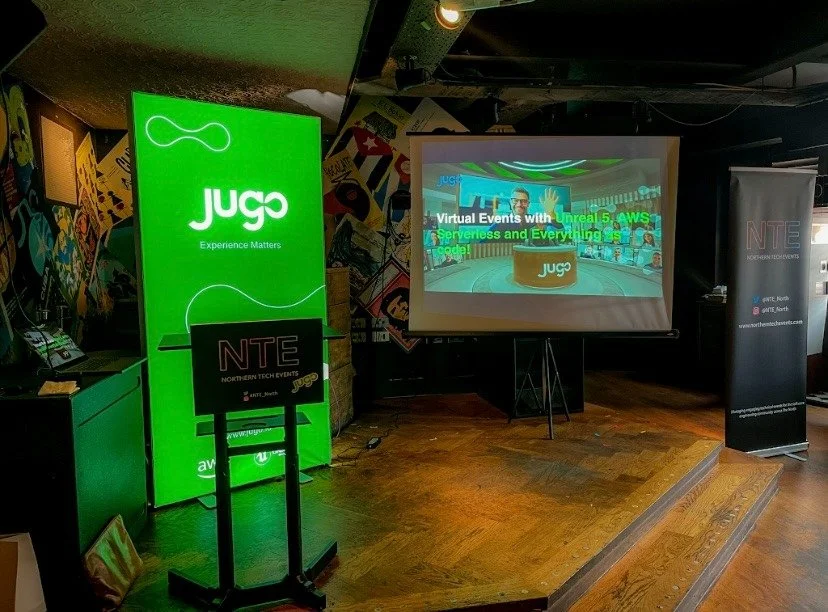 Event setup with a large illuminated green sign displaying the 'Jugo' logo and slogan, a projection screen showing a virtual event titled 'Virtual Events with Unreal 5, AWS, Serverless and Everything in between,' a small black stand with the 'NTE' logo, and a black banner with the NTE logo on the right, all on a wooden floor with a comic-style mural on the wall background.