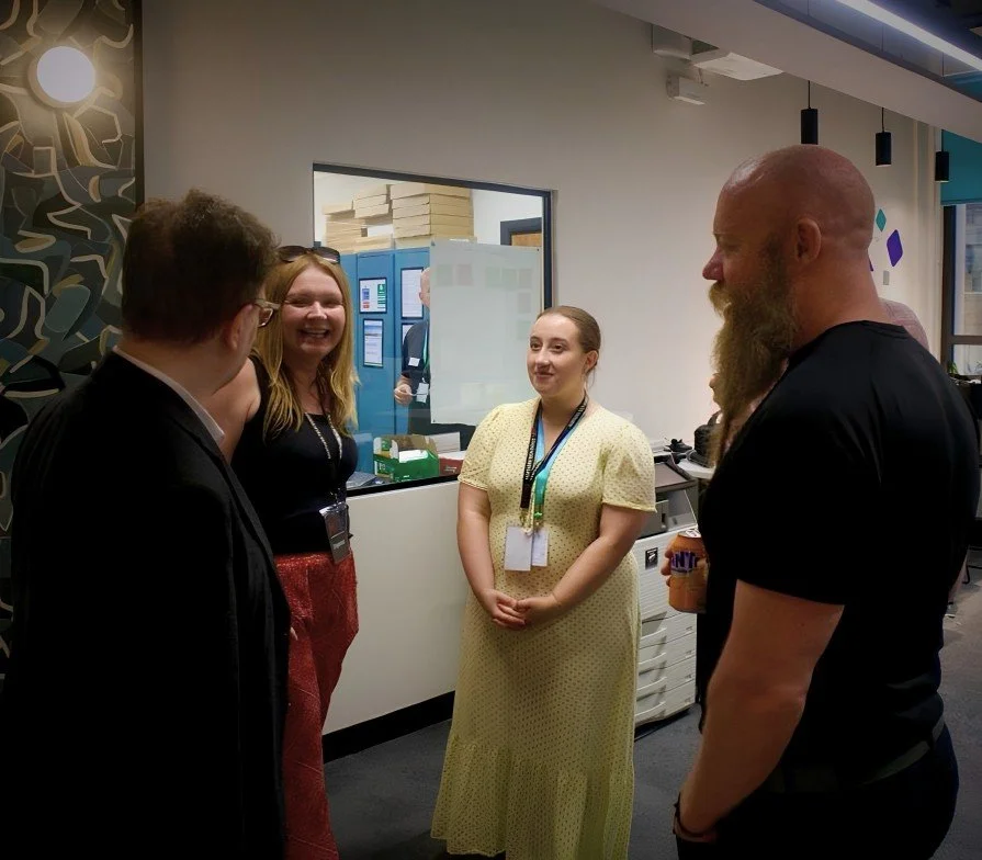 Four people having a conversation in an office, one person is holding a cup, and two others are wearing lanyards.