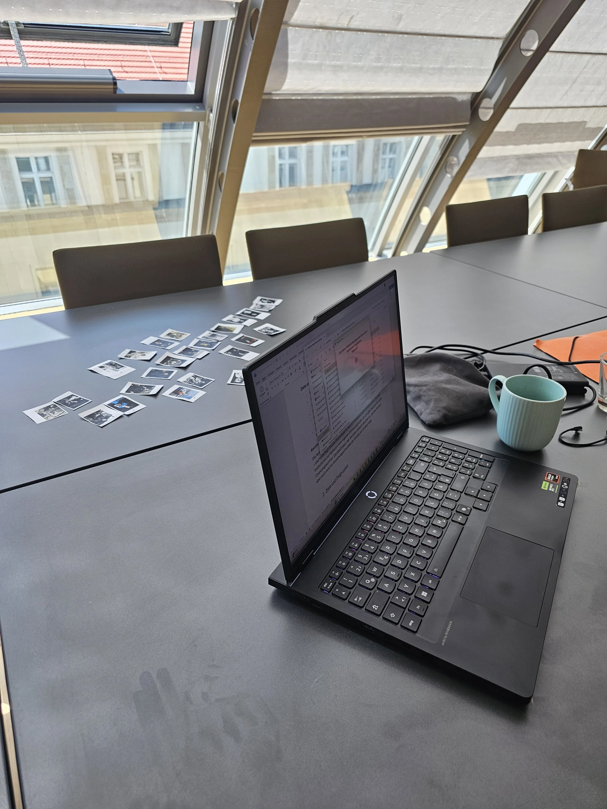 Ein Schreibtisch im Konferenzraum mit einem Laptop, einer Tasse, einem Notizbuch und einer Reihe von kleineren Fotos auf der Oberfläche.