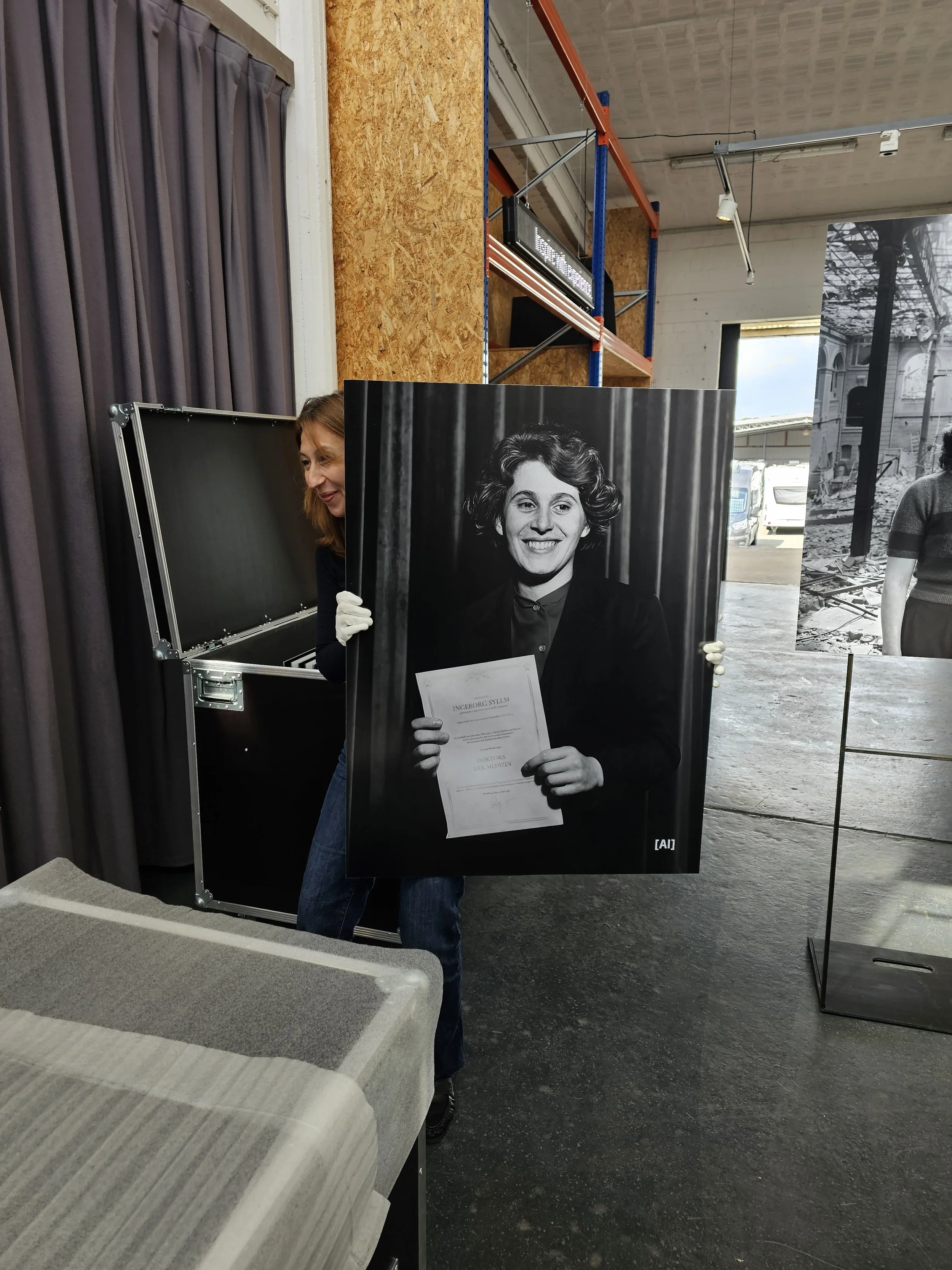 Eine Frau mit roten Haaren richtet sich hinter einem großen Schwarz-Weiß-Foto einer lachenden Frau mit kurzen, gewellten Haaren auf. Sie trägt eine dunkle Jacke und hält das Foto mit beiden Händen. Es scheint sich um eine Ausstellung oder eine Präsentation zu handeln.
