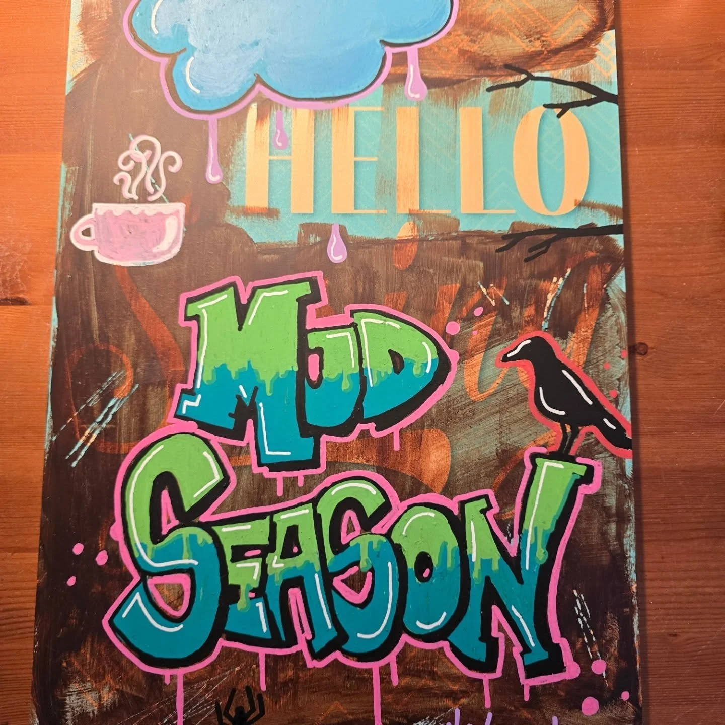 Colorful graffiti art with the words 'HELLO SEASON' in bold, vibrant letters, a pink steaming cup, a blue cloud with rain, a black bird, and a spider on a branch, on a wooden surface.