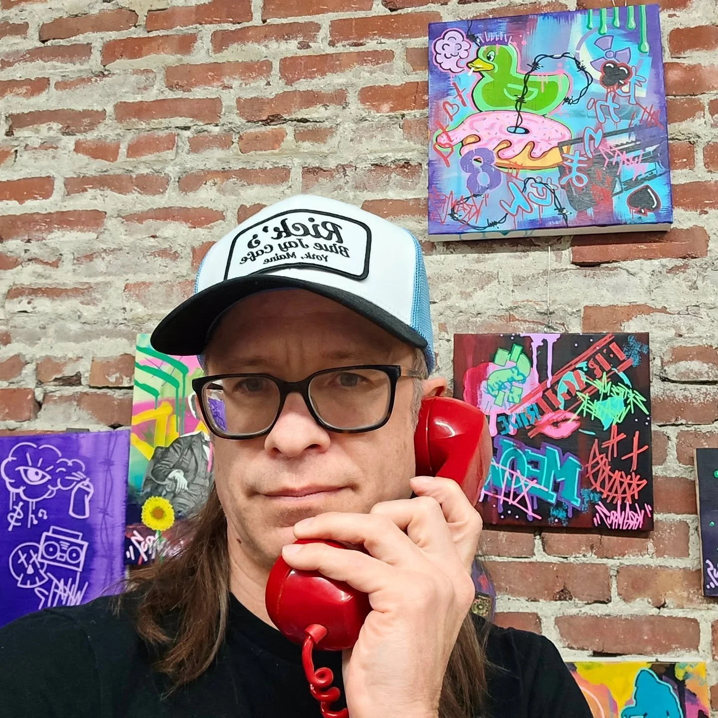 A man with long hair, glasses, wearing a white and black trucker hat, holding a red vintage rotary phone to his ear. Behind him is a brick wall decorated with colorful graffiti-style artwork.