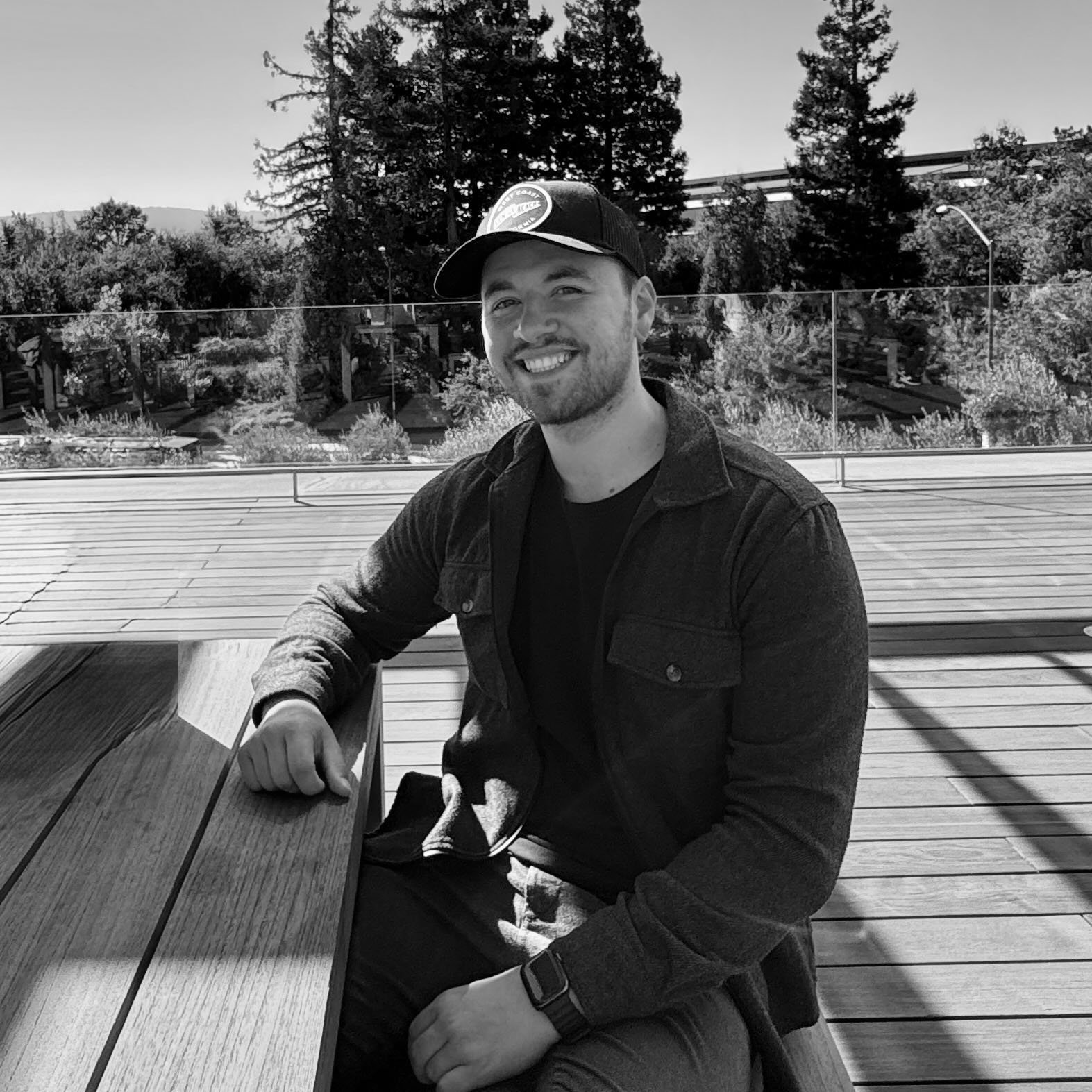 Ein lächelnder junger Mann in dunkler Kleidung sitzt an einem holzverkleideten Tisch im Freien, im Hintergrund sind Bäume und eine Glasabsperrung. Es ist eine sonnige Tageszeit.