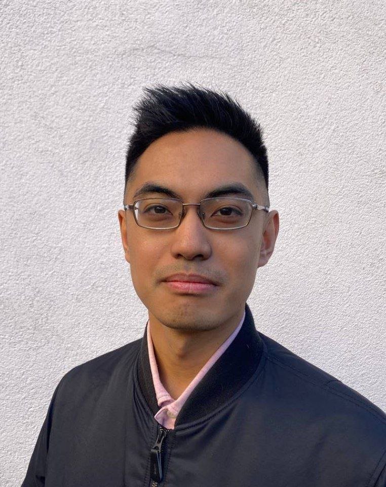 A young Asian man with short black hair and glasses wearing a black jacket over a pink collared shirt, standing against a plain white wall.