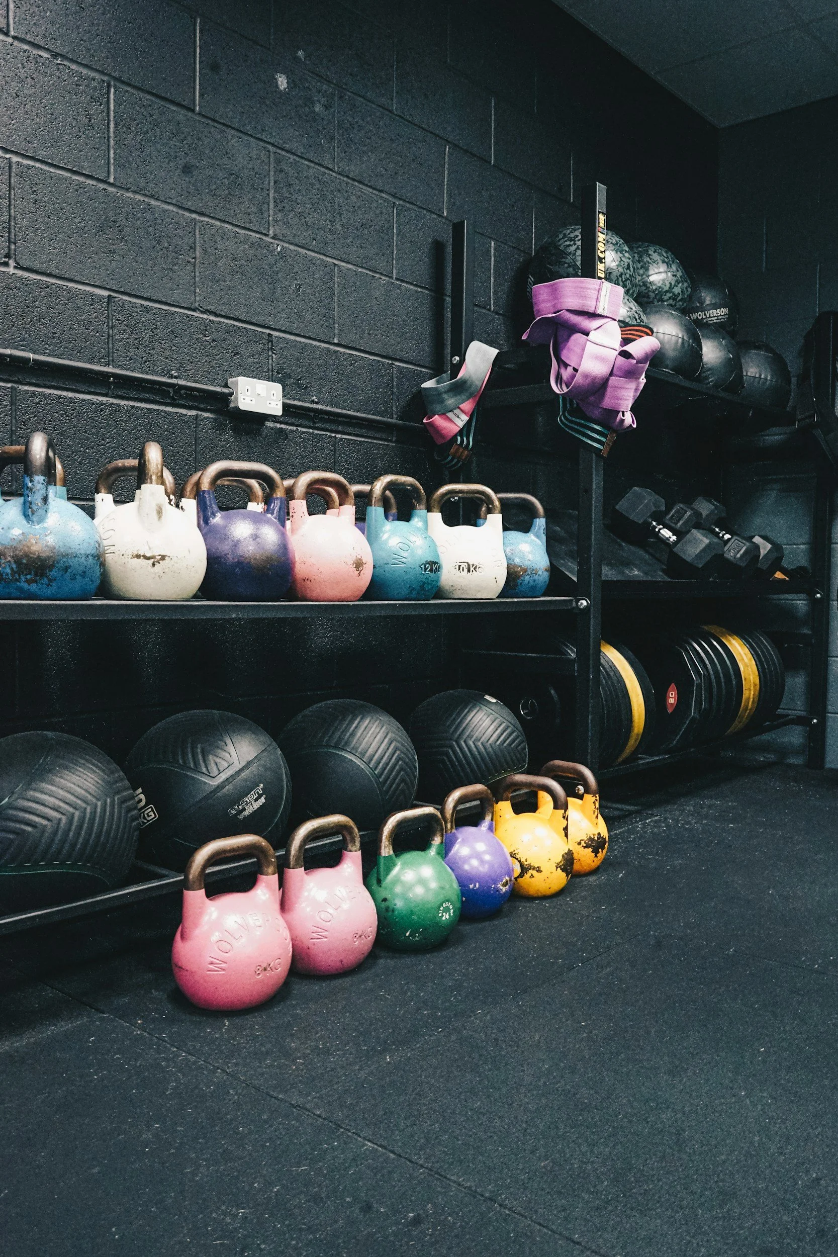 Kettlebells, medicine balls, and resistance bands stored on black metal shelves in a gym with black brick walls.
