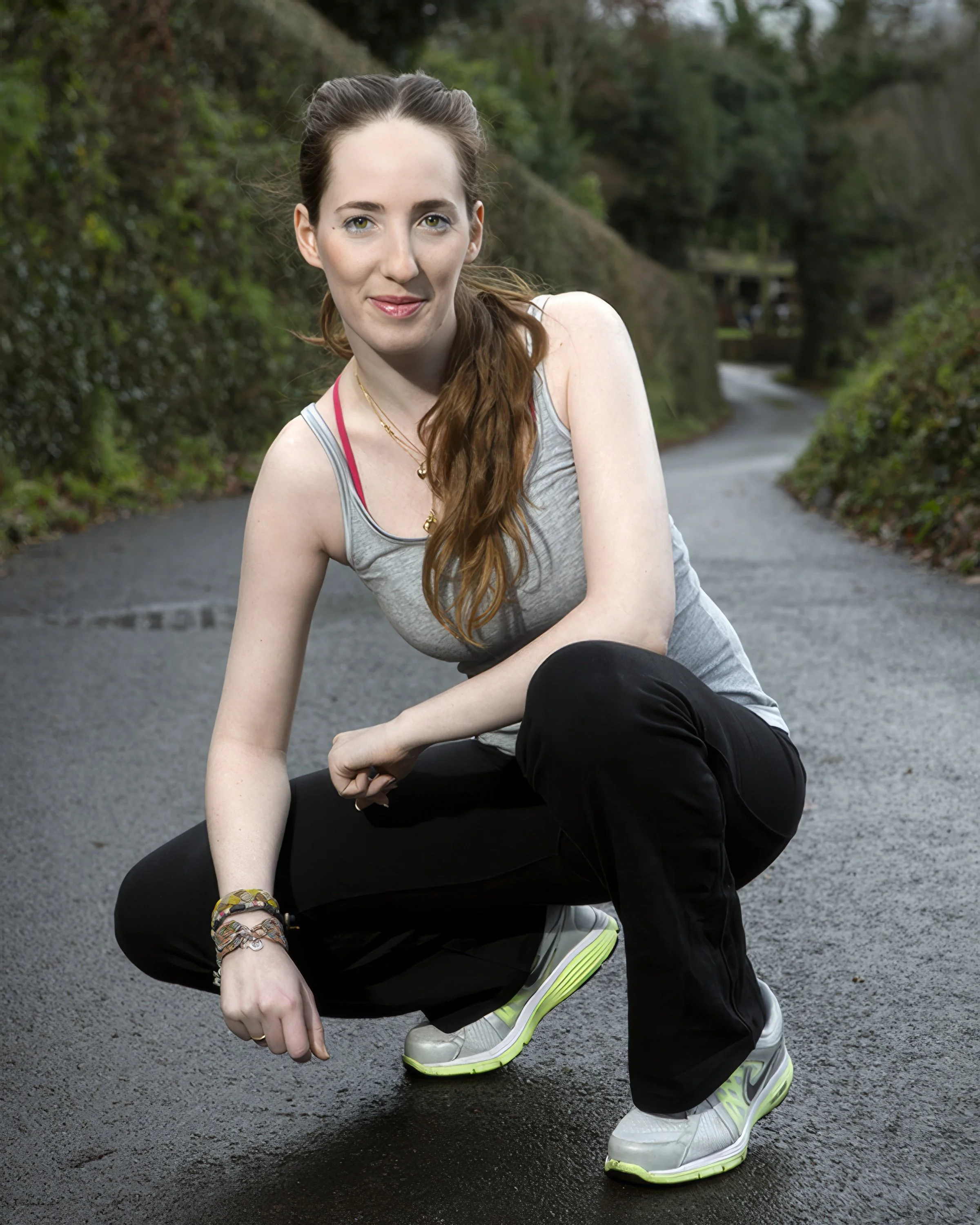 A young woman with long brown hair in a ponytail, wearing a gray tank top with pink straps, black pants, and white sneakers with lime green soles, posed outdoors on a paved path surrounded by trees and greenery.