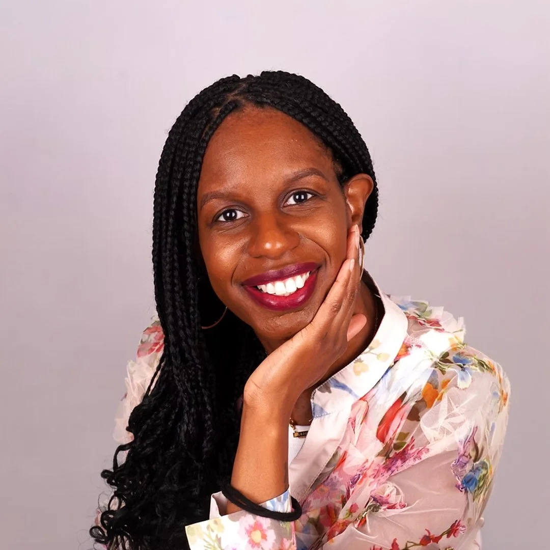 Portrait of a woman with braided hair smiling and resting her face on her hand, wearing a floral blouse.
