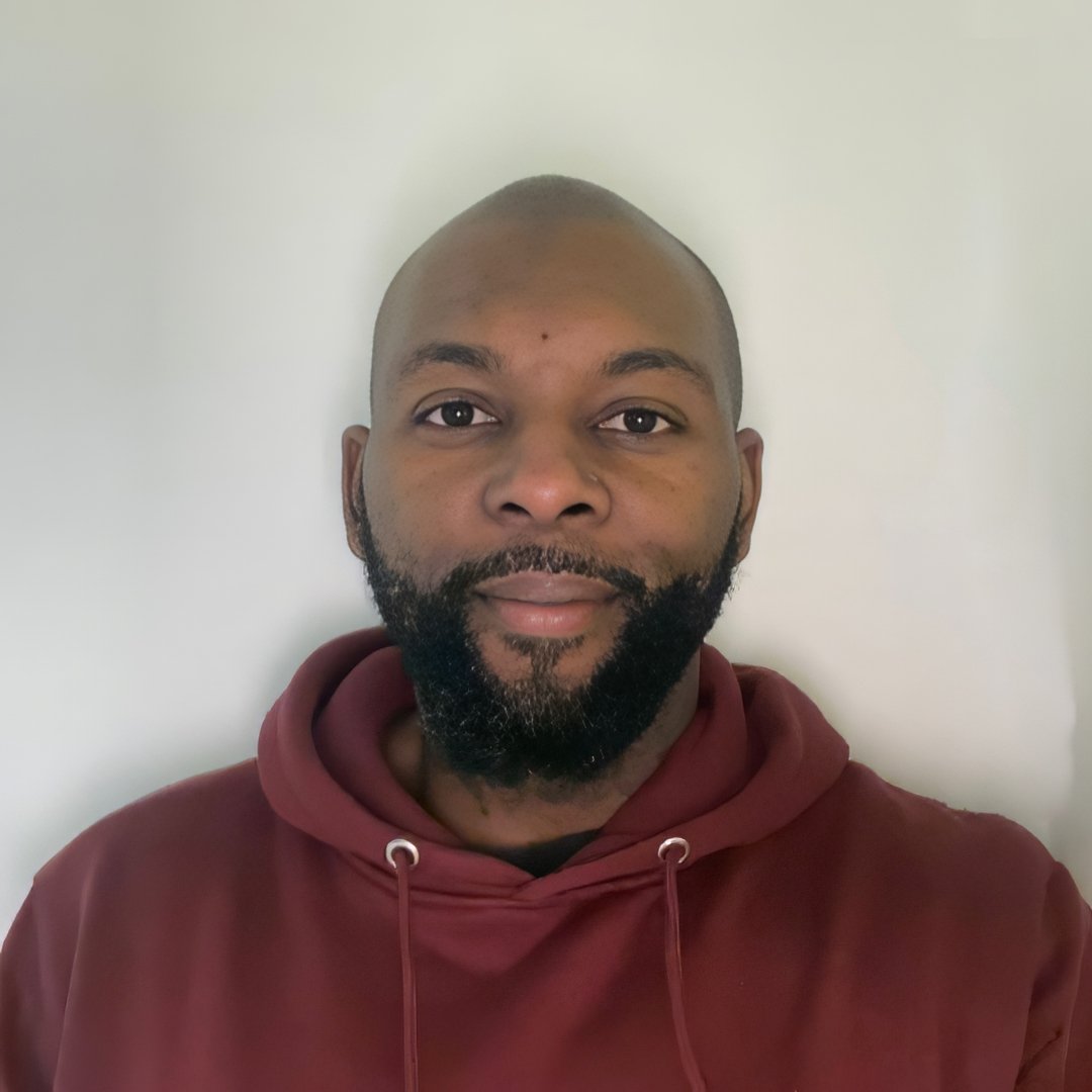 A man with a beard and shaved head wearing a maroon hoodie standing against a light-colored wall.