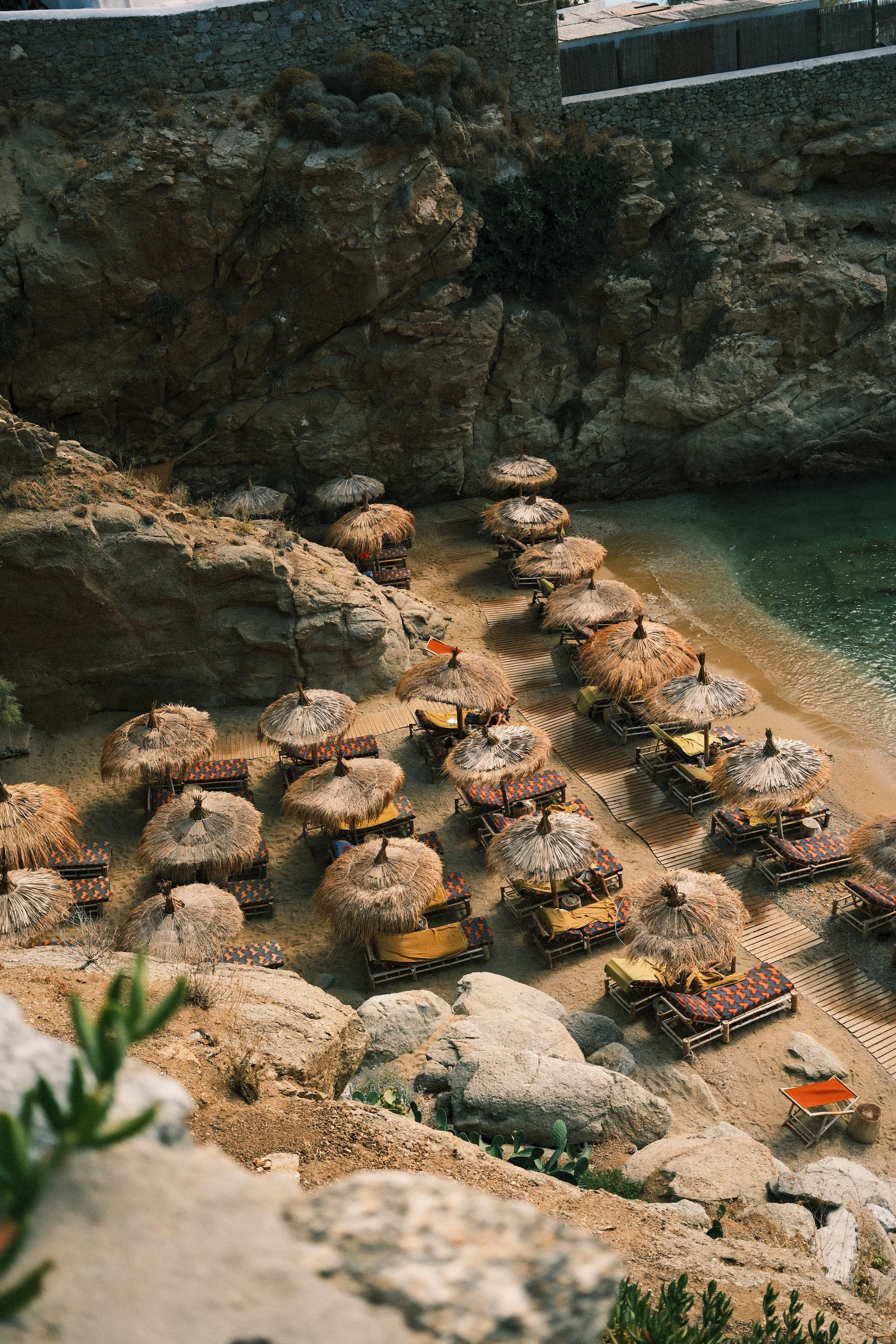 A beach scene with numerous straw umbrellas and lounge chairs along the shoreline, with rocky cliffs in the background and a small section of water visible on the right side.