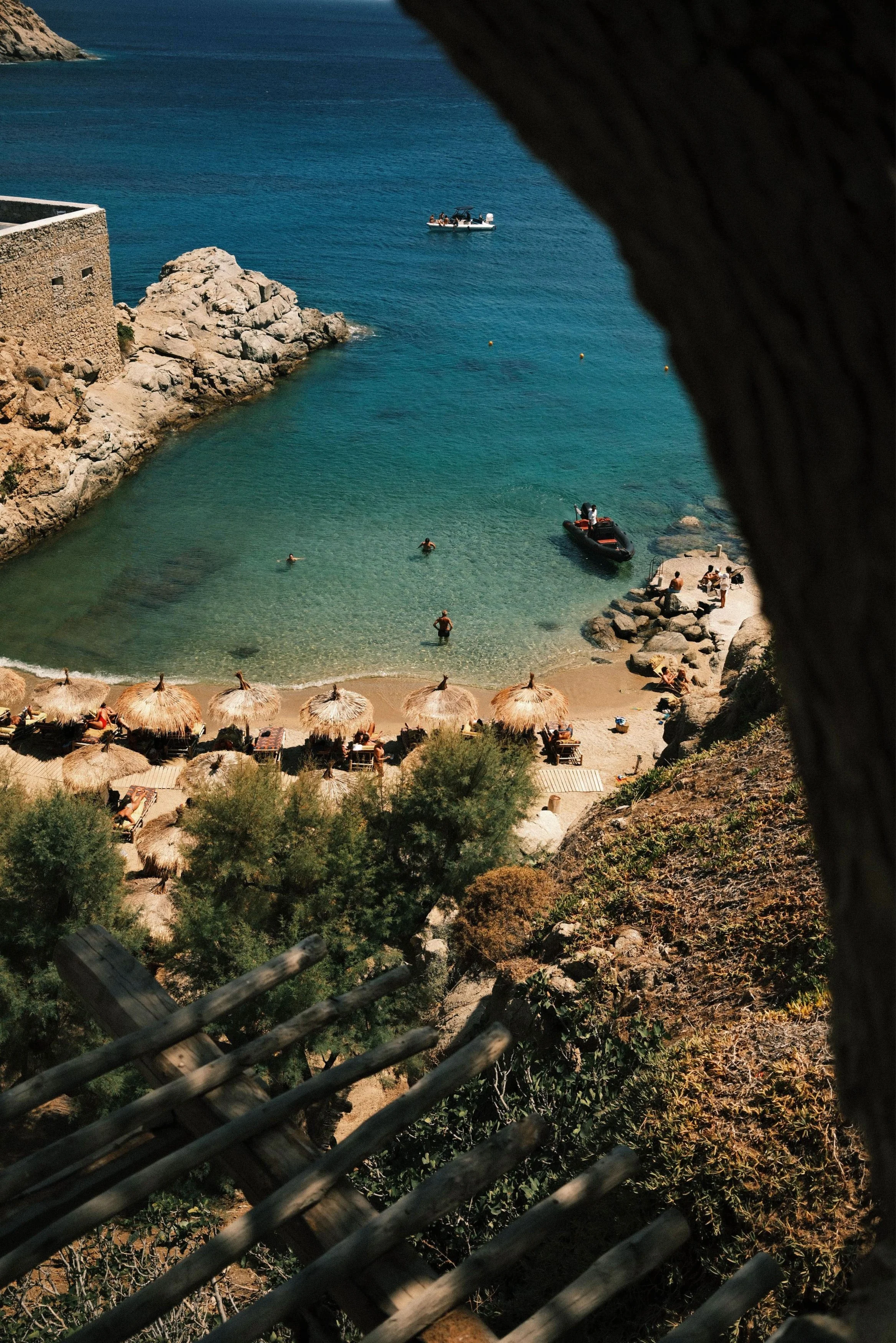 A beach scene viewed through a wooden fence and a natural arch or rock formation. The beach has straw umbrellas and people swimming, with clear water and rocky coastline. A boat floats on the water.