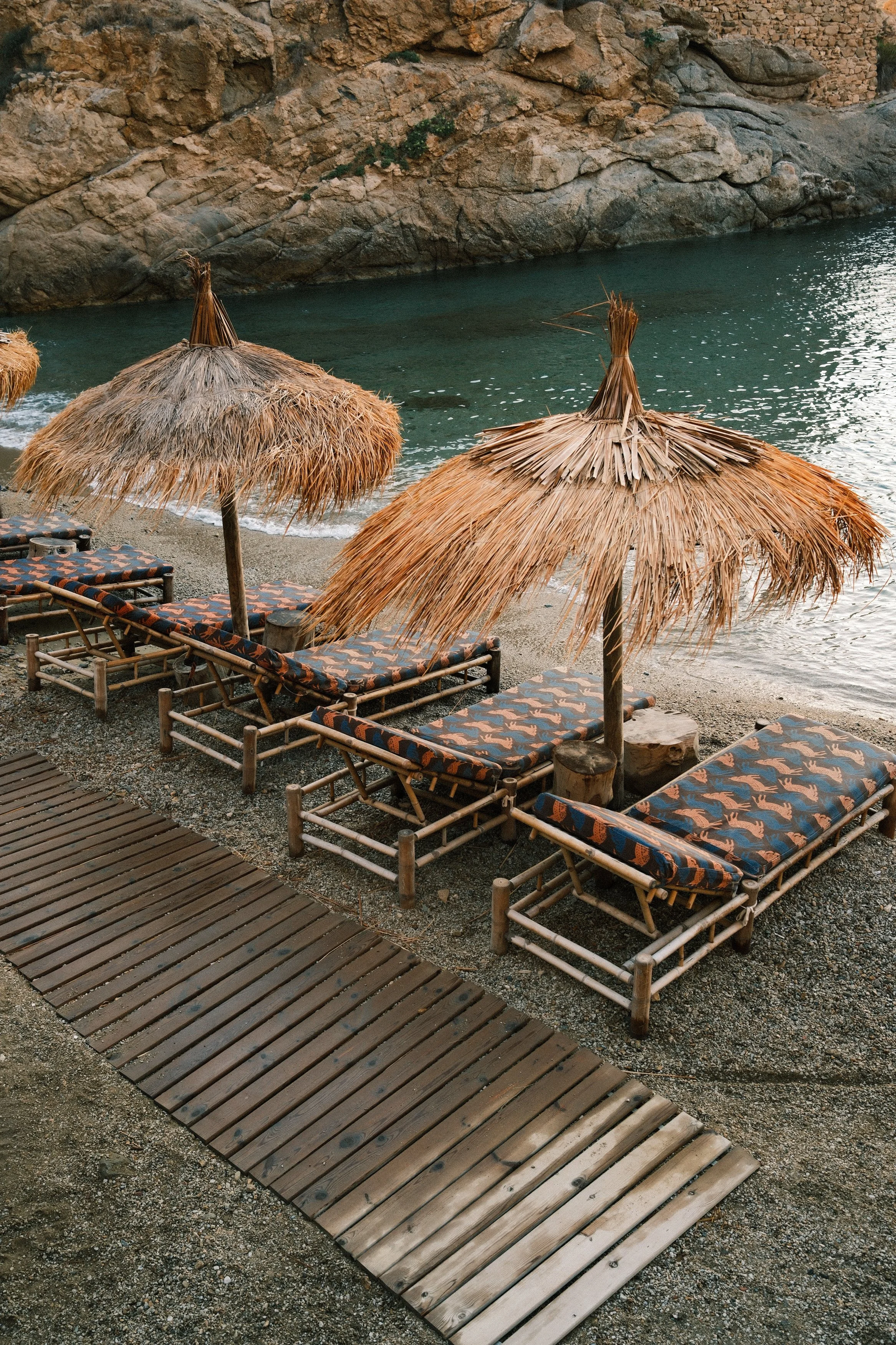 Beach scene with straw umbrellas and lounge chairs on sand, rocky cliff in the background, and a wooden pathway in the foreground.