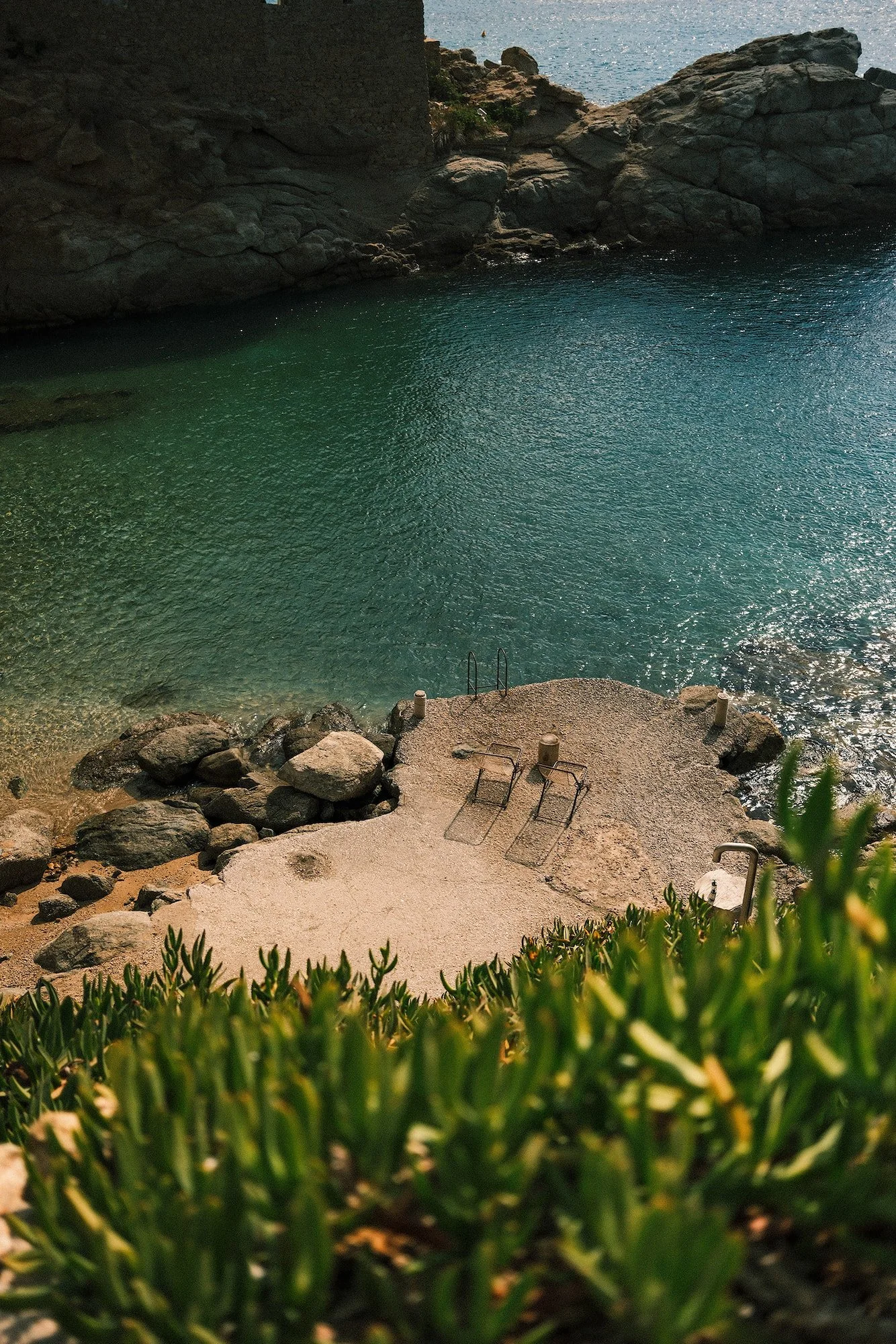 A secluded beach with a concrete platform, four empty lounge chairs, surrounded by rocks and greenery, overlooking calm turquoise water.