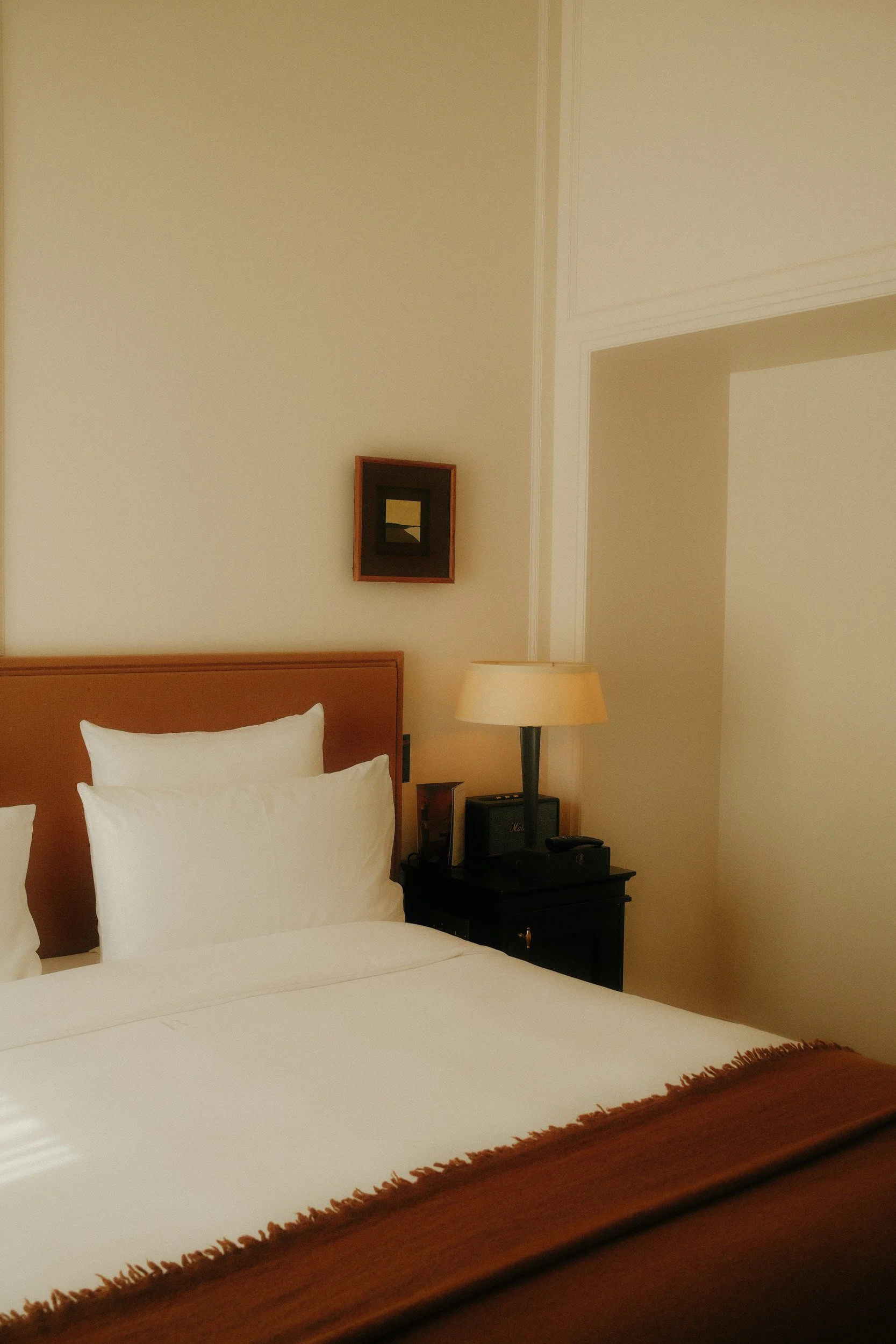 A hotel room with a bed, white pillows, a bedside lamp, a small wooden nightstand, and a framed picture on a beige wall.