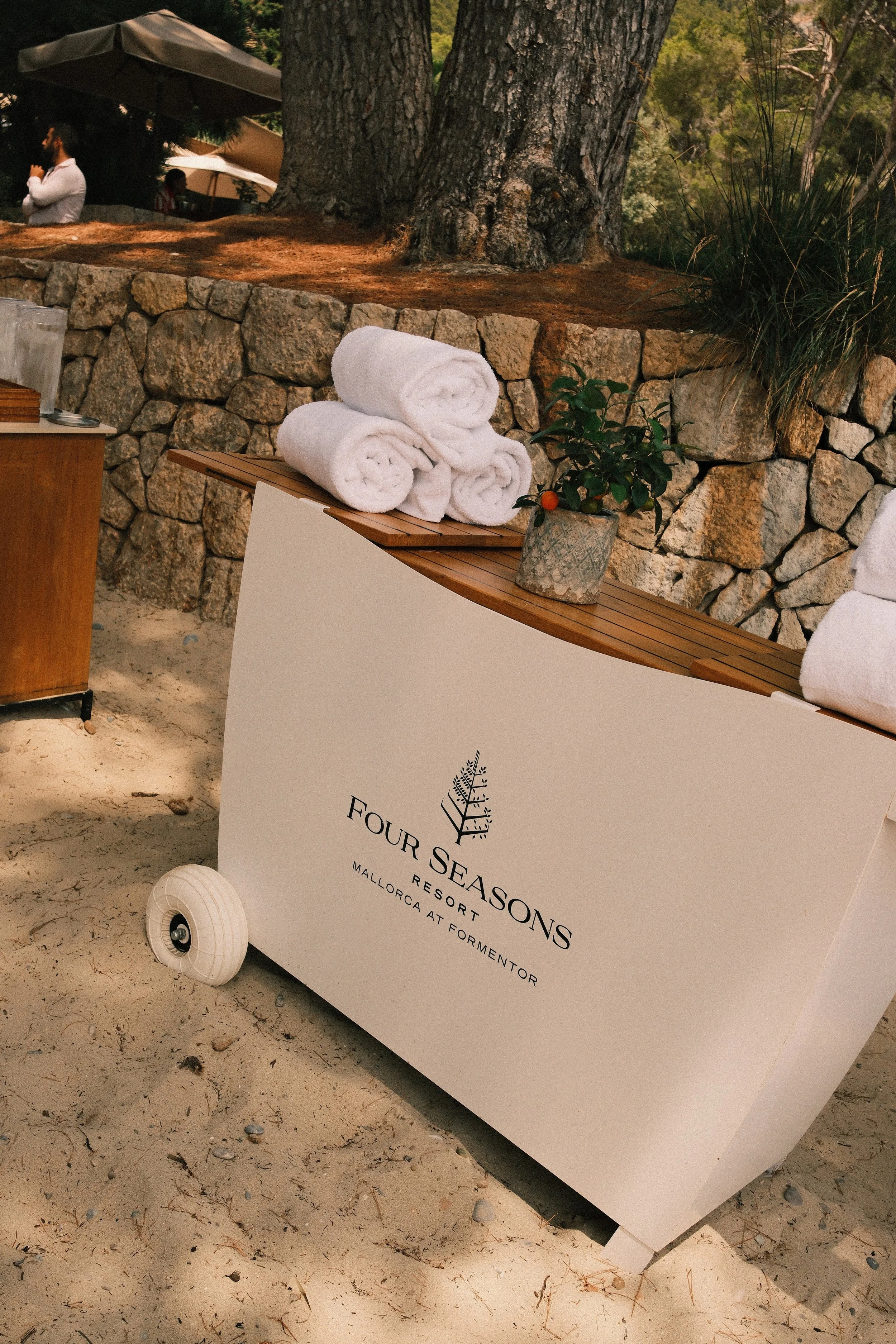 Stack of three rolled white towels on a wooden table with a potted plant beside them, set outdoors in a natural setting with trees and a stone wall in the background.