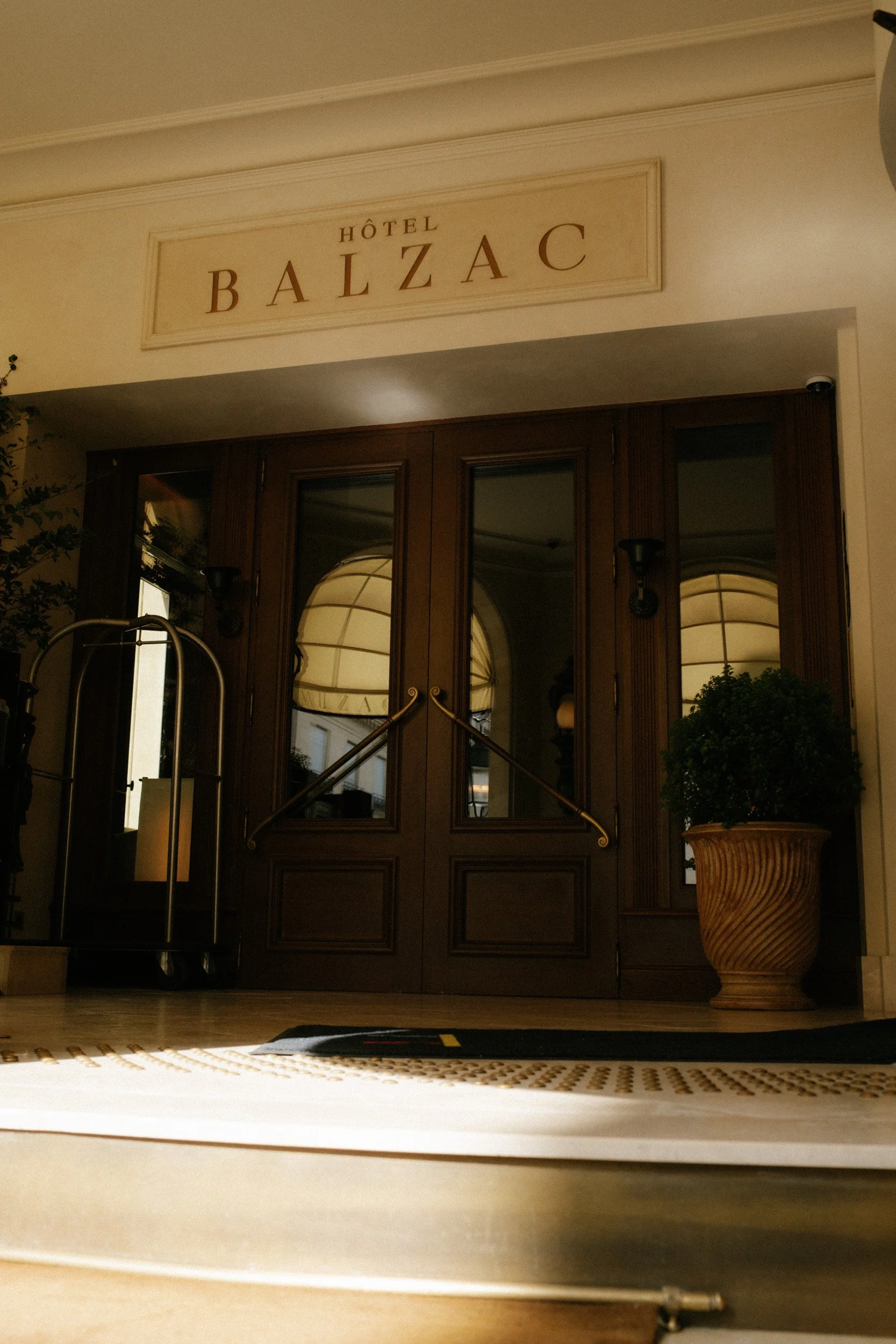 Entrance of a hotel named Hotel Balzac with wooden double doors and large potted plant on the right side.